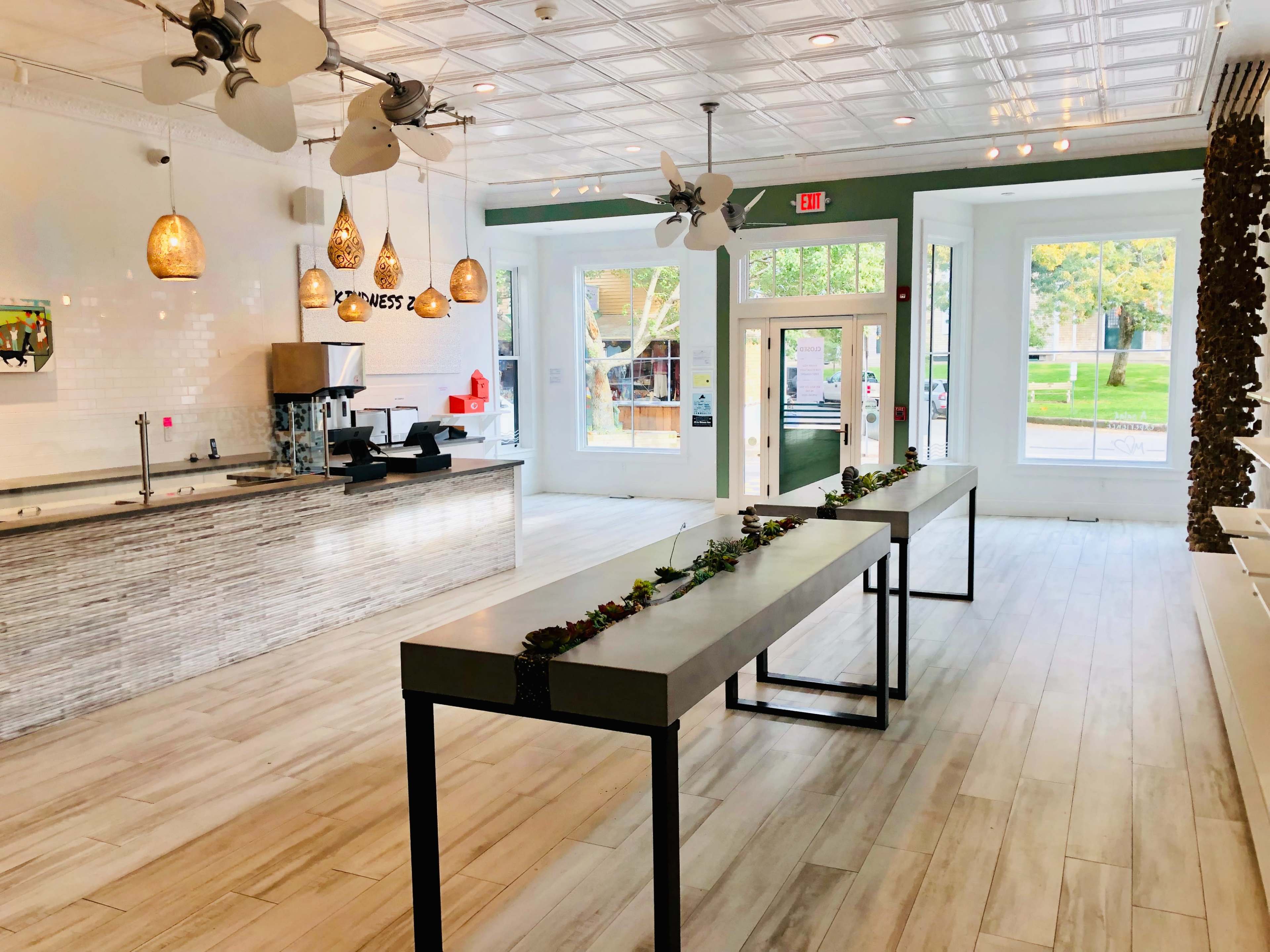 A modern, airy café interior featuring a counter with a coffee machine and pendant lights, two large communal tables, and windows that provide natural light.