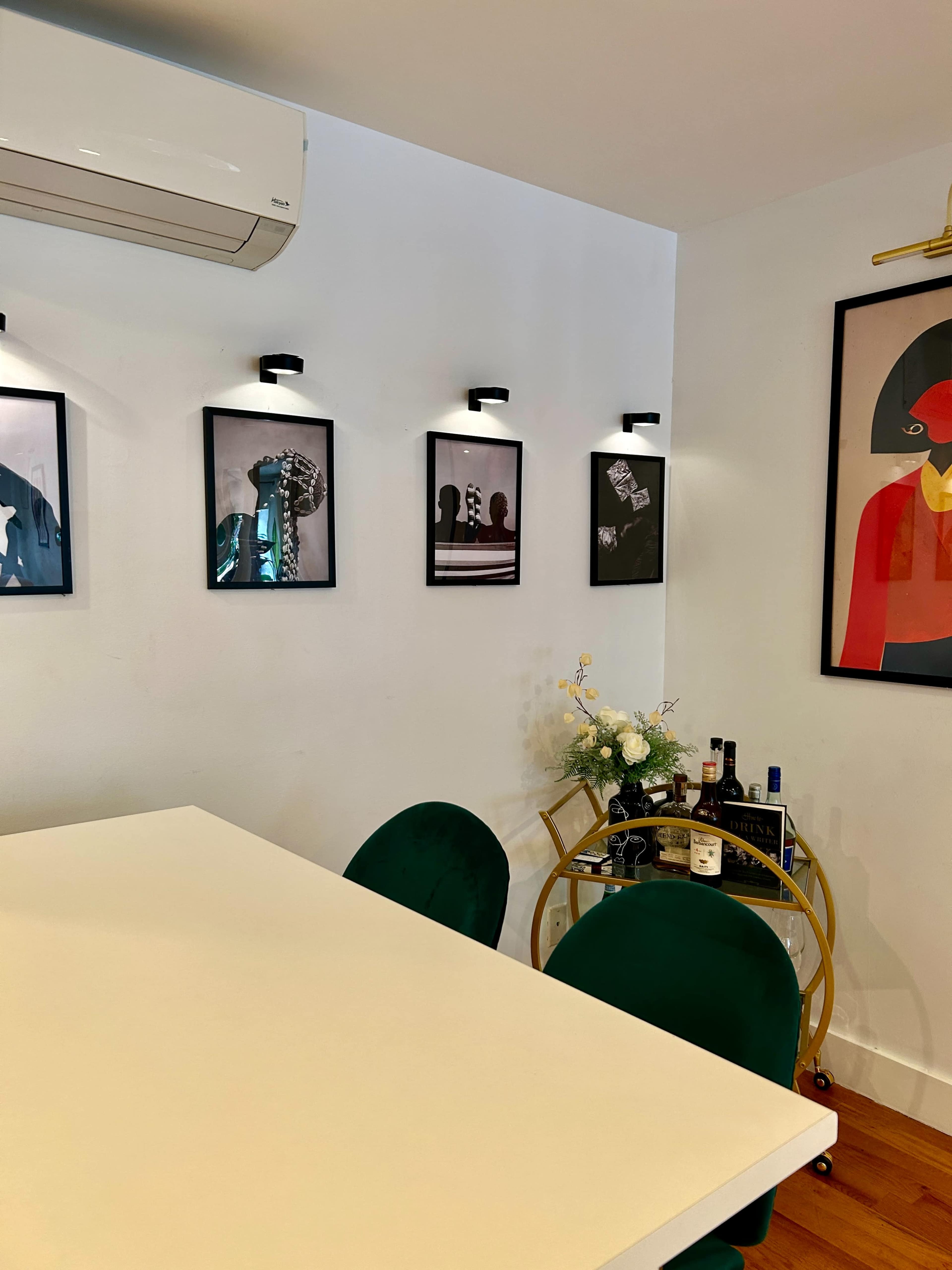 A modern indoor space featuring a white table with green chairs, decorated walls with framed artworks, and a bar cart with bottles and flowers.