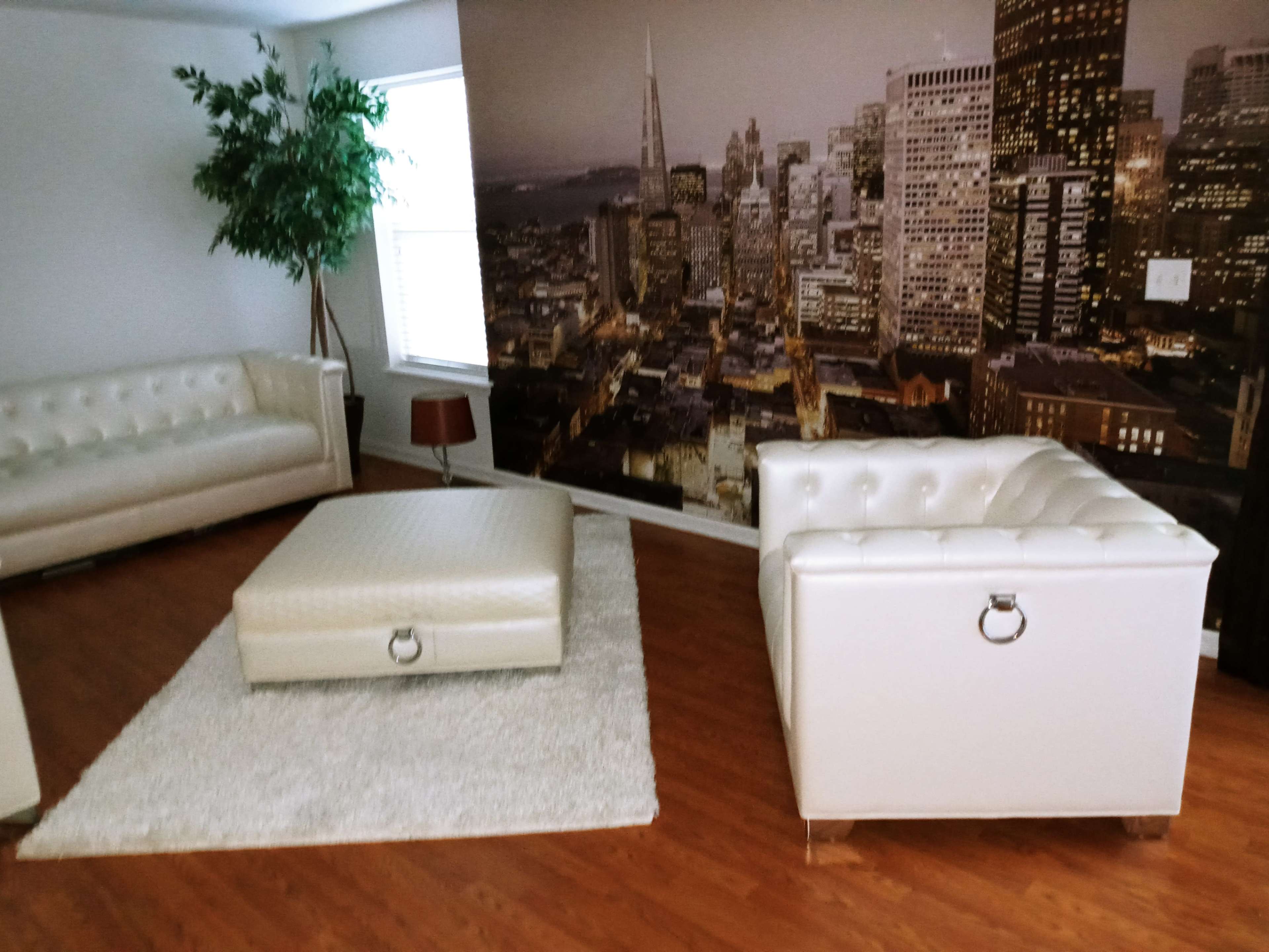 The scene features a modern living area with two white tufted sofas, an ottoman, a small round lamp, and a wall mural depicting a city skyline.