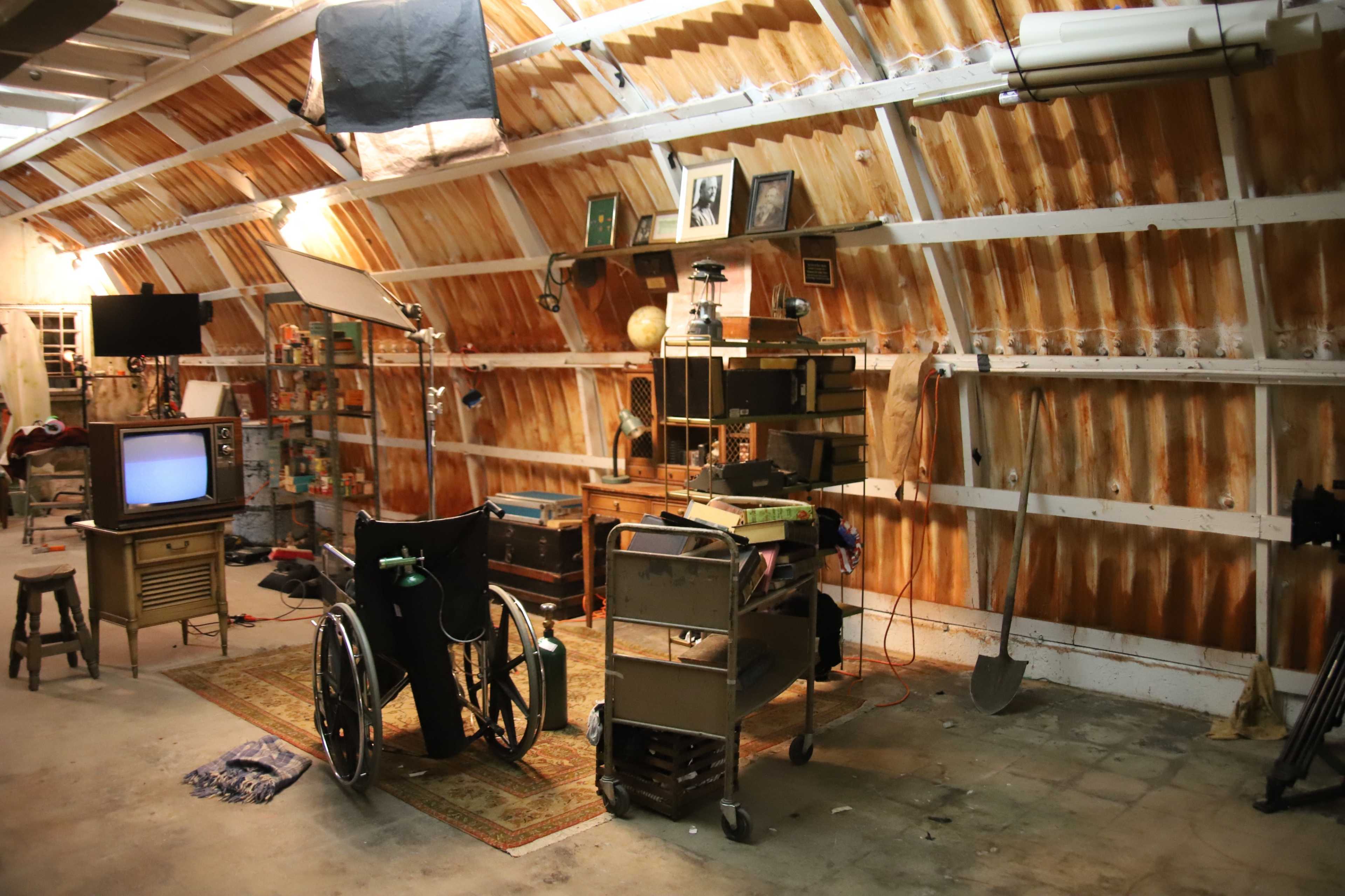 A spacious garage with wooden walls, featuring a vintage television, a wheelchair, and several cluttered worktables and tools.