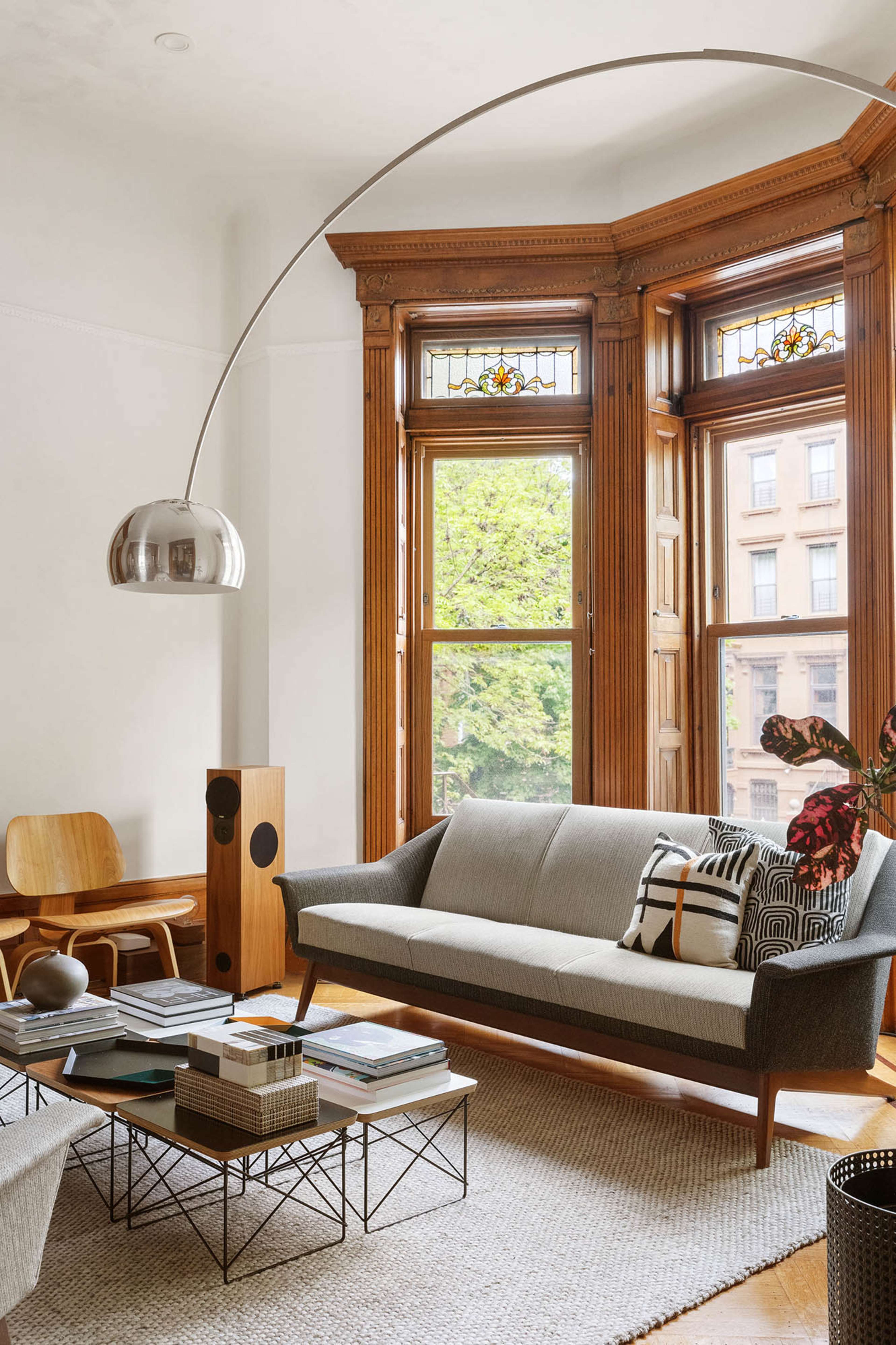 A modern living room features a gray sofa, a large arc floor lamp, and wooden accents with large windows overlooking a tree-lined street.