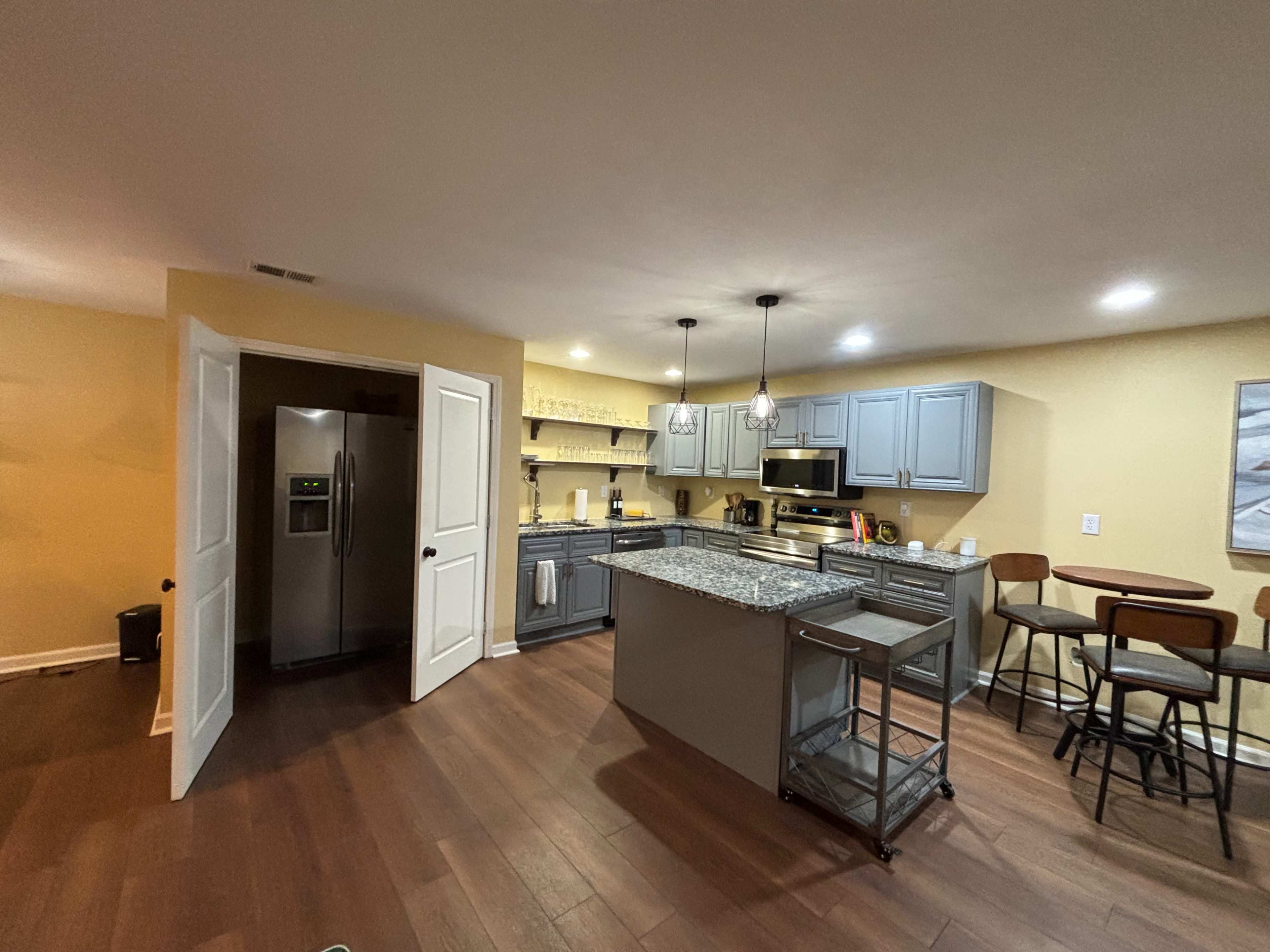 The image shows a modern kitchen with gray cabinets, a large island, stainless steel appliances, and a dining table with stools nearby.