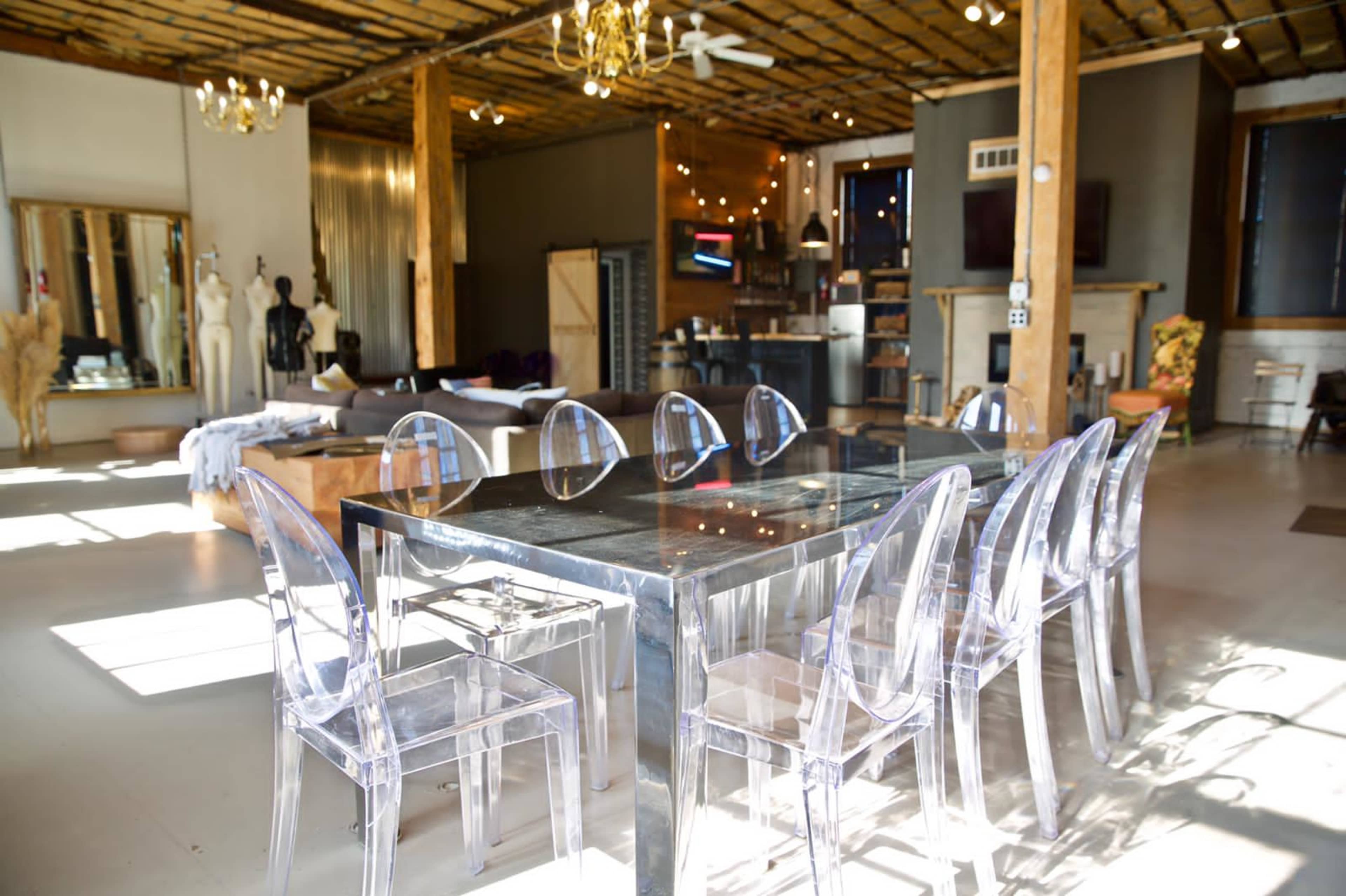 The image shows a spacious interior featuring a modern dining table with transparent chairs, wooden beams, and various decorative elements.