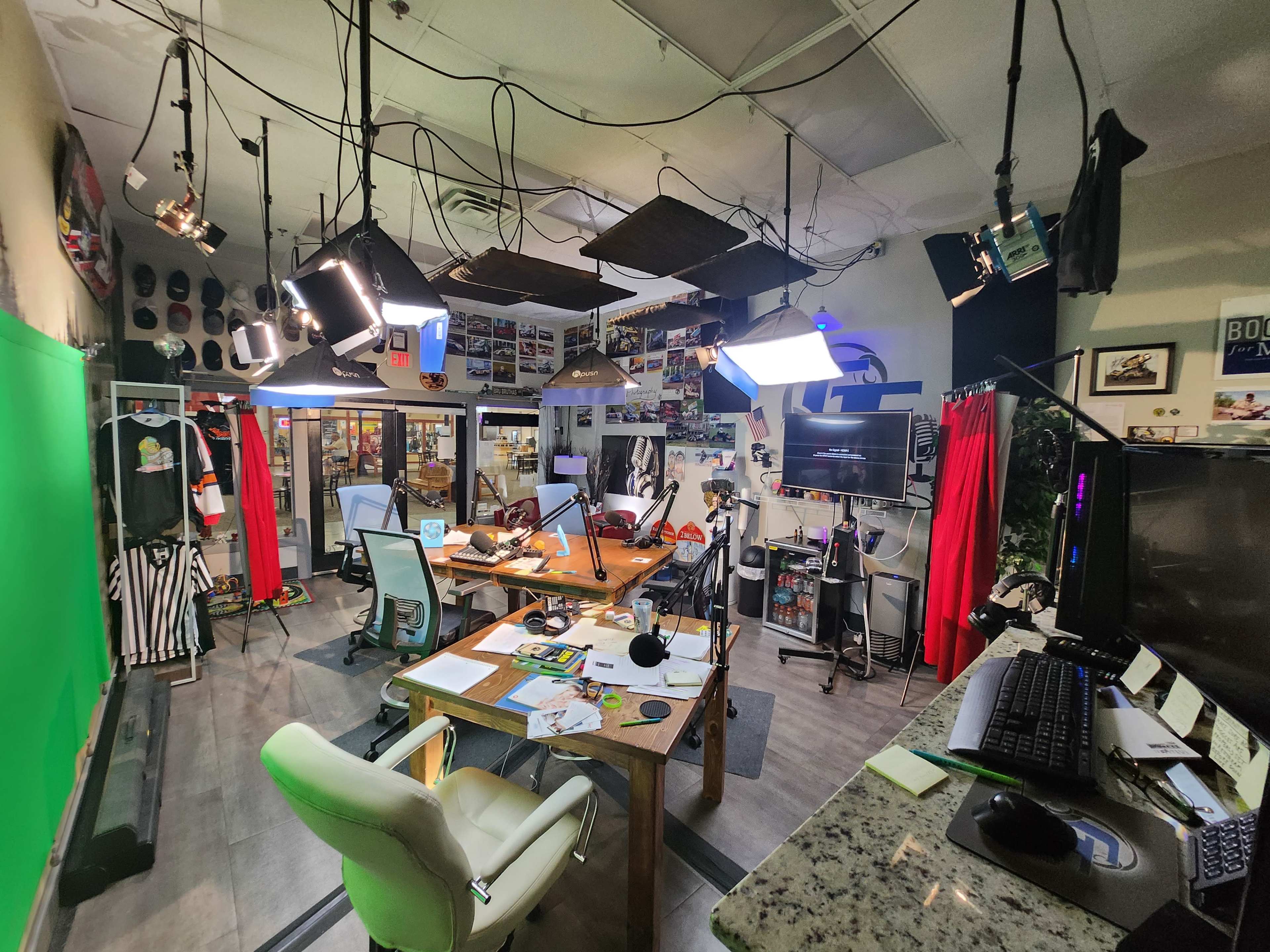A production studio with a green screen, various lighting setups, desks, and a wall decorated with photographs and memorabilia.
