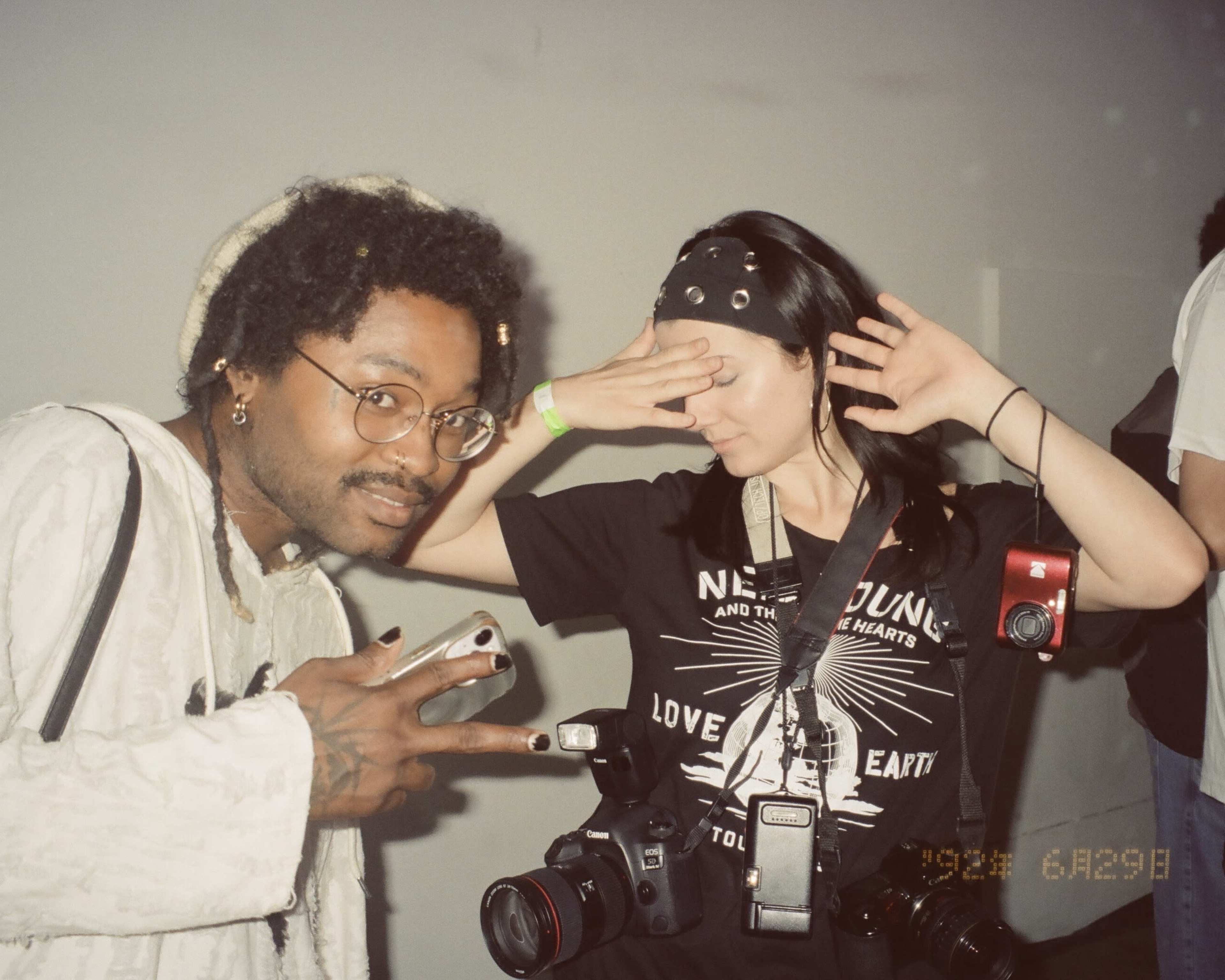 A man and a woman pose for a photo, with the man holding up his phone and making a peace sign, while the woman stands with her hand over her forehead and multiple cameras hanging around her neck.
