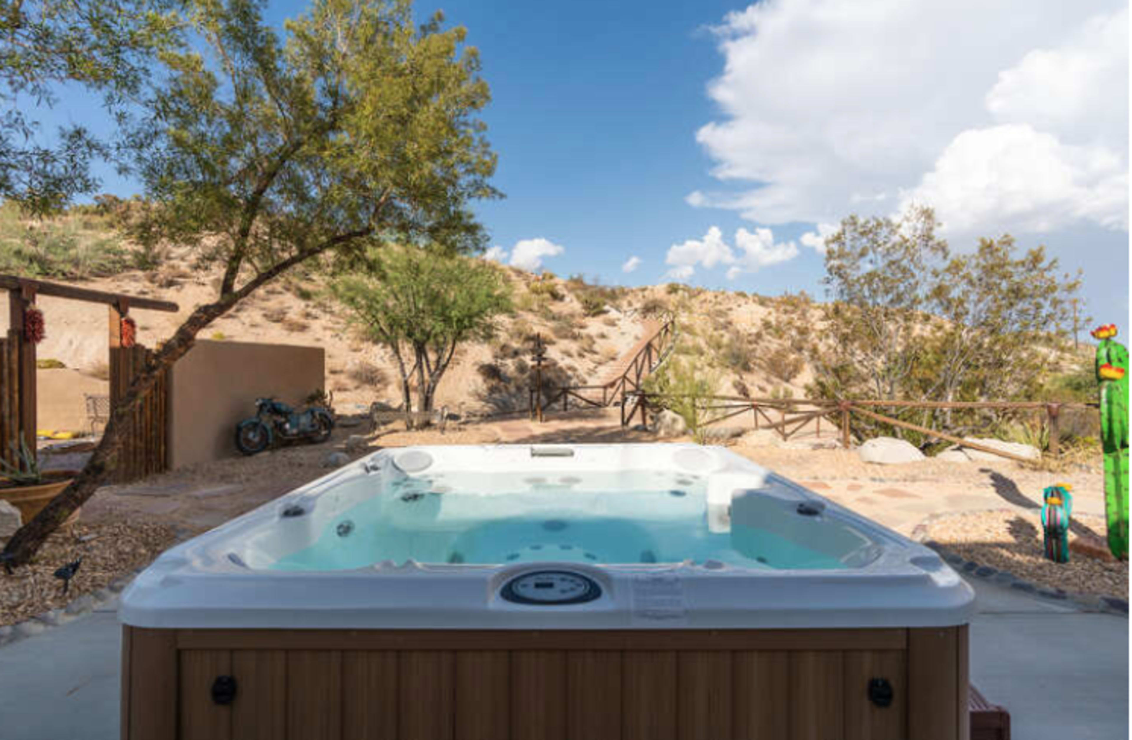 A hot tub sits on a patio surrounded by desert scenery, with trees and a mountain in the background.