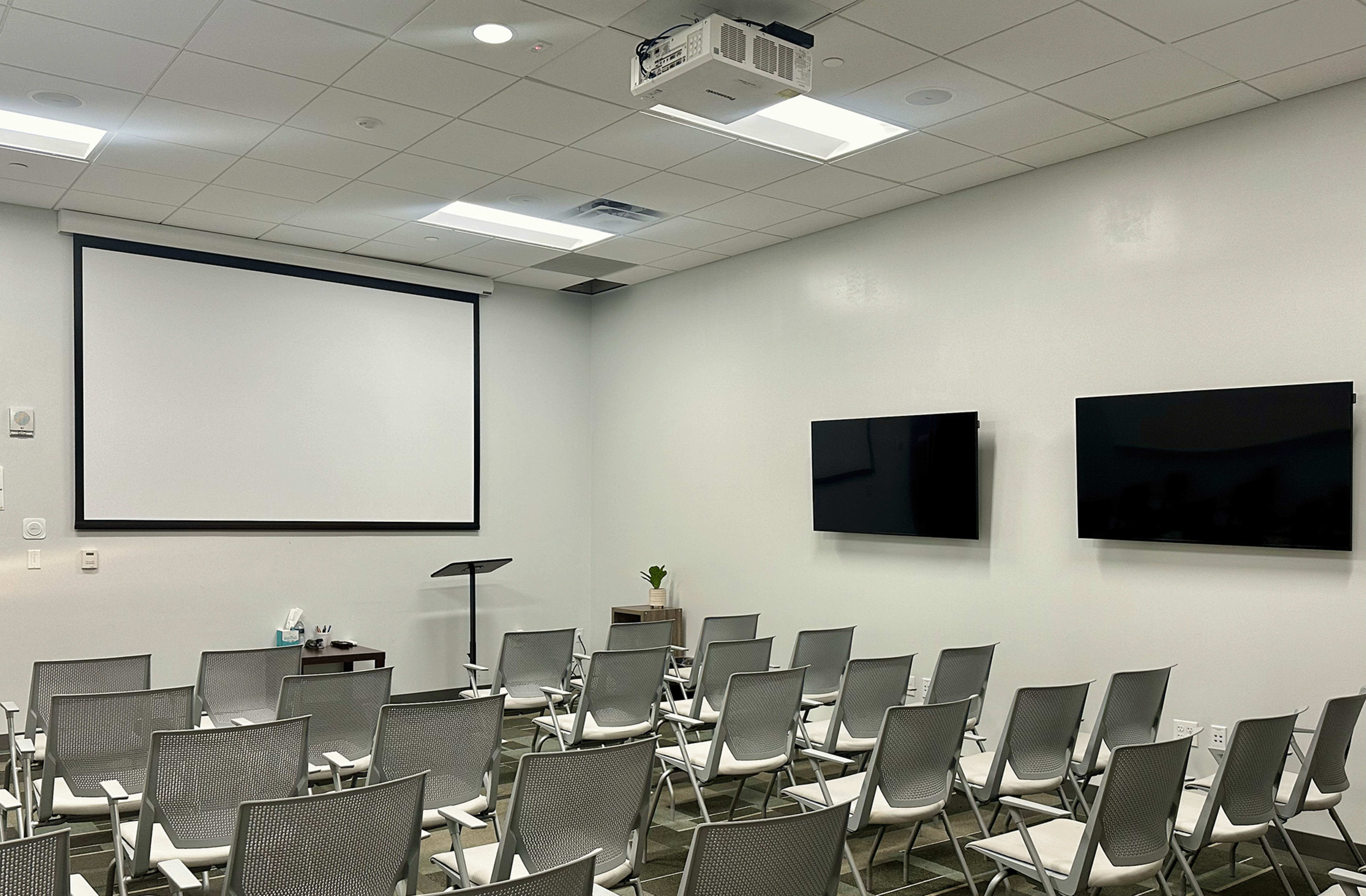 A modern meeting room with rows of silver chairs facing a projector screen and two mounted televisions on the wall.