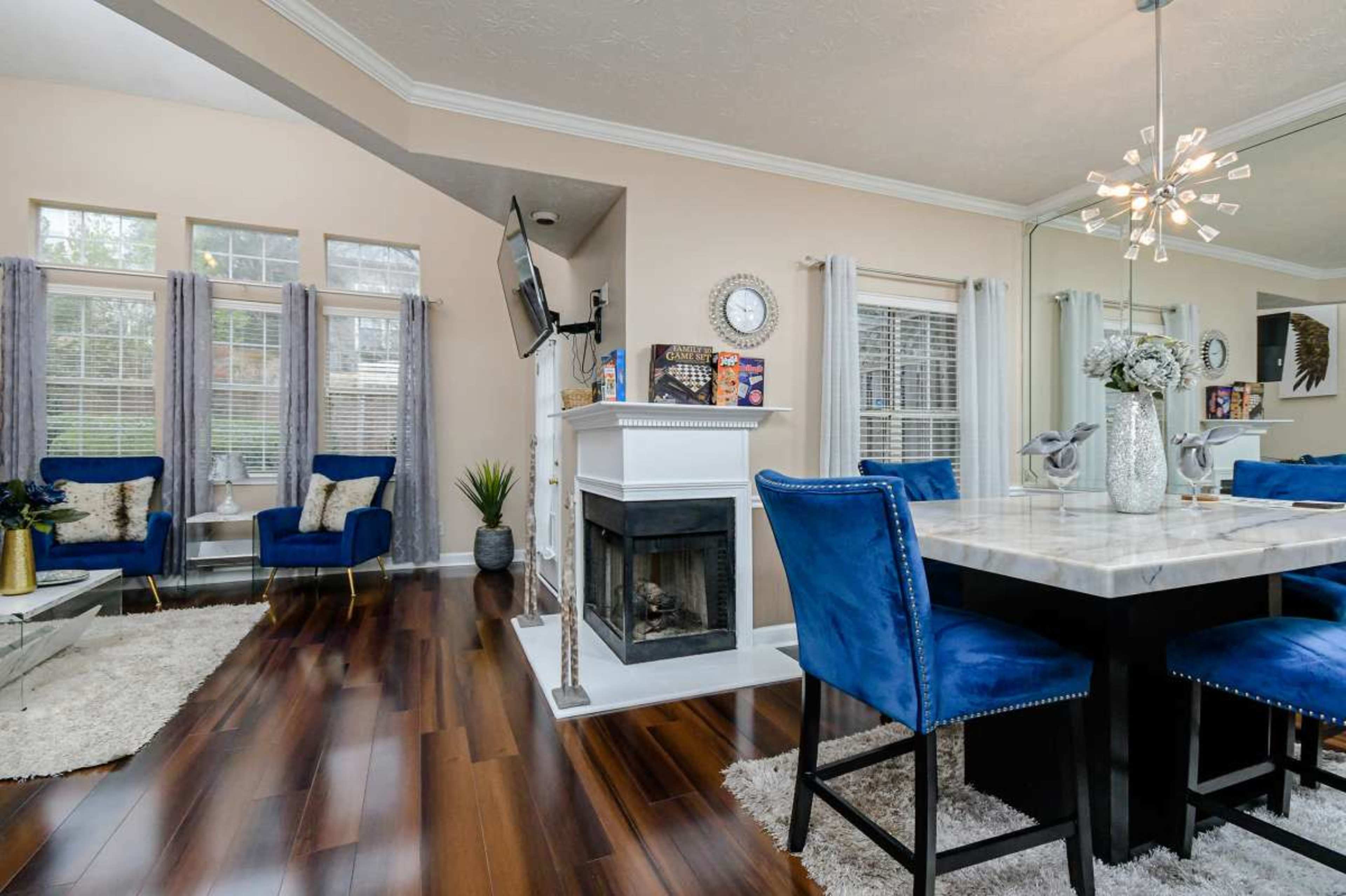 The image shows a modern interior featuring a dining area with a marble table and blue upholstered chairs, alongside a living space with a fireplace and decorative shelving.