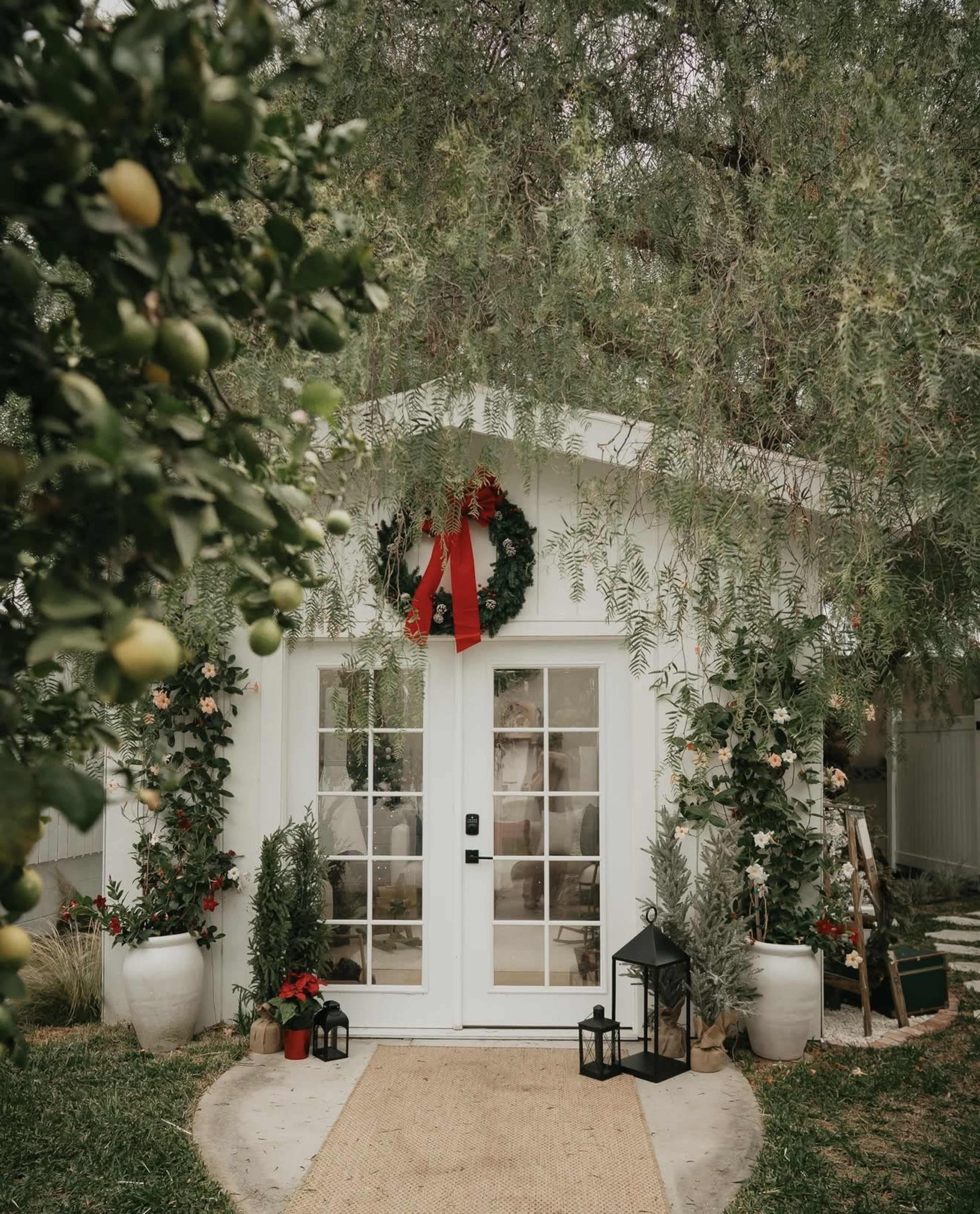 A light-colored cottage entrance is adorned with a green wreath and red bow, surrounded by potted plants and lanterns, set against a backdrop of lush greenery.