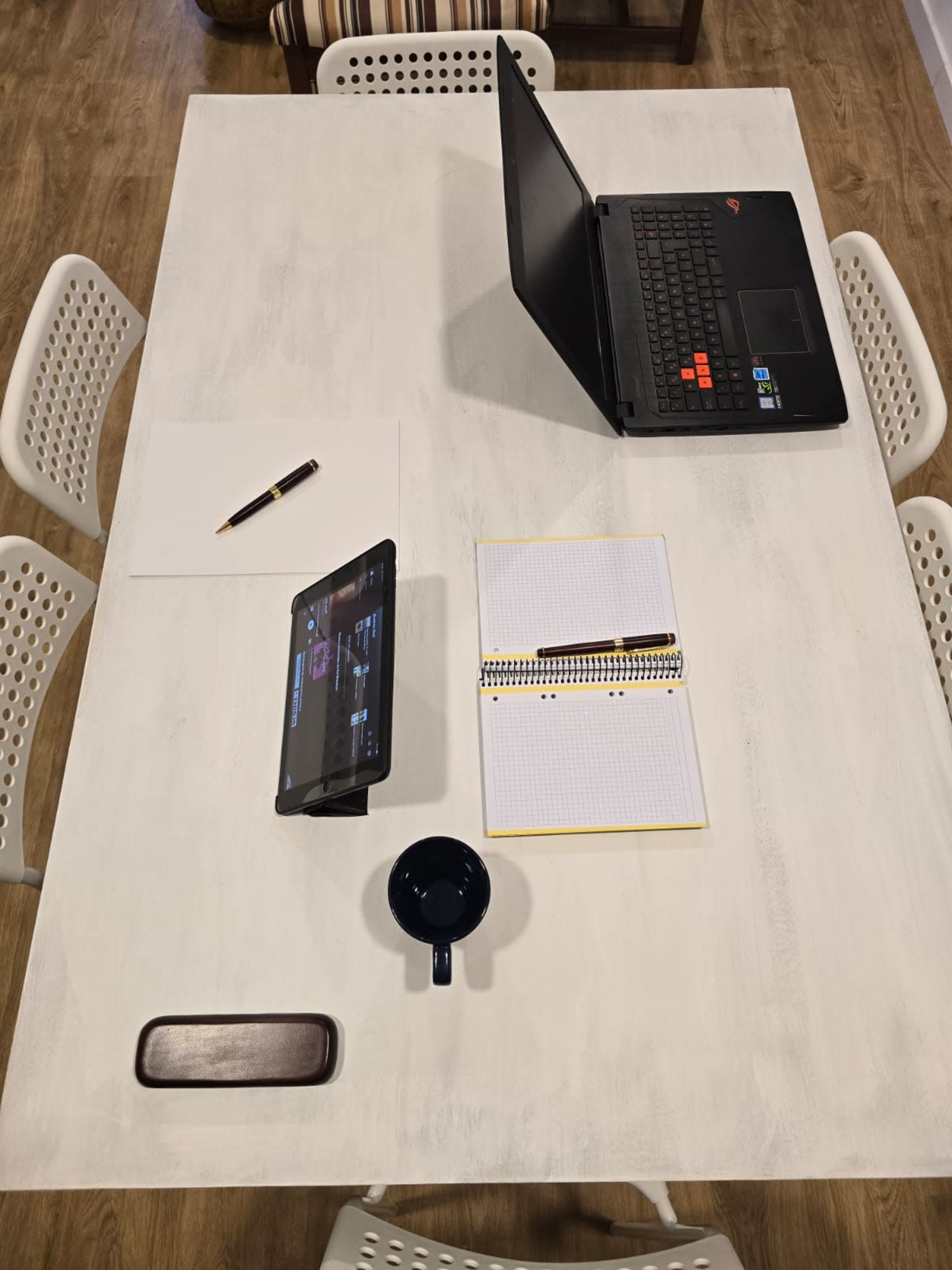 A laptop, tablet, notebook, pen, and mug are positioned on a white table with a sheet of paper.