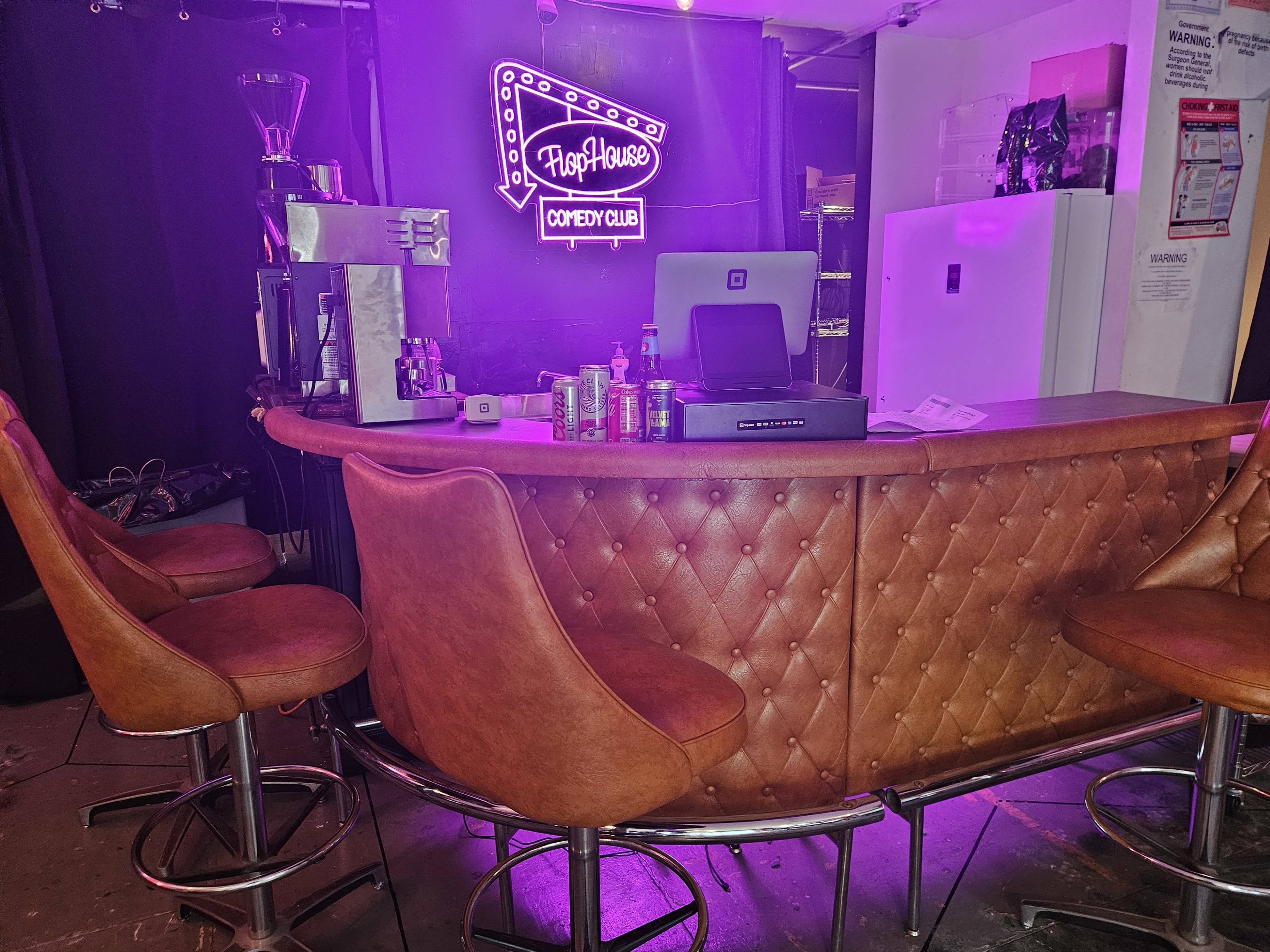 The image shows a cozy bar area with a brown leather counter, several high stools, and a purple neon sign that reads "FlopHouse Comedy Club."