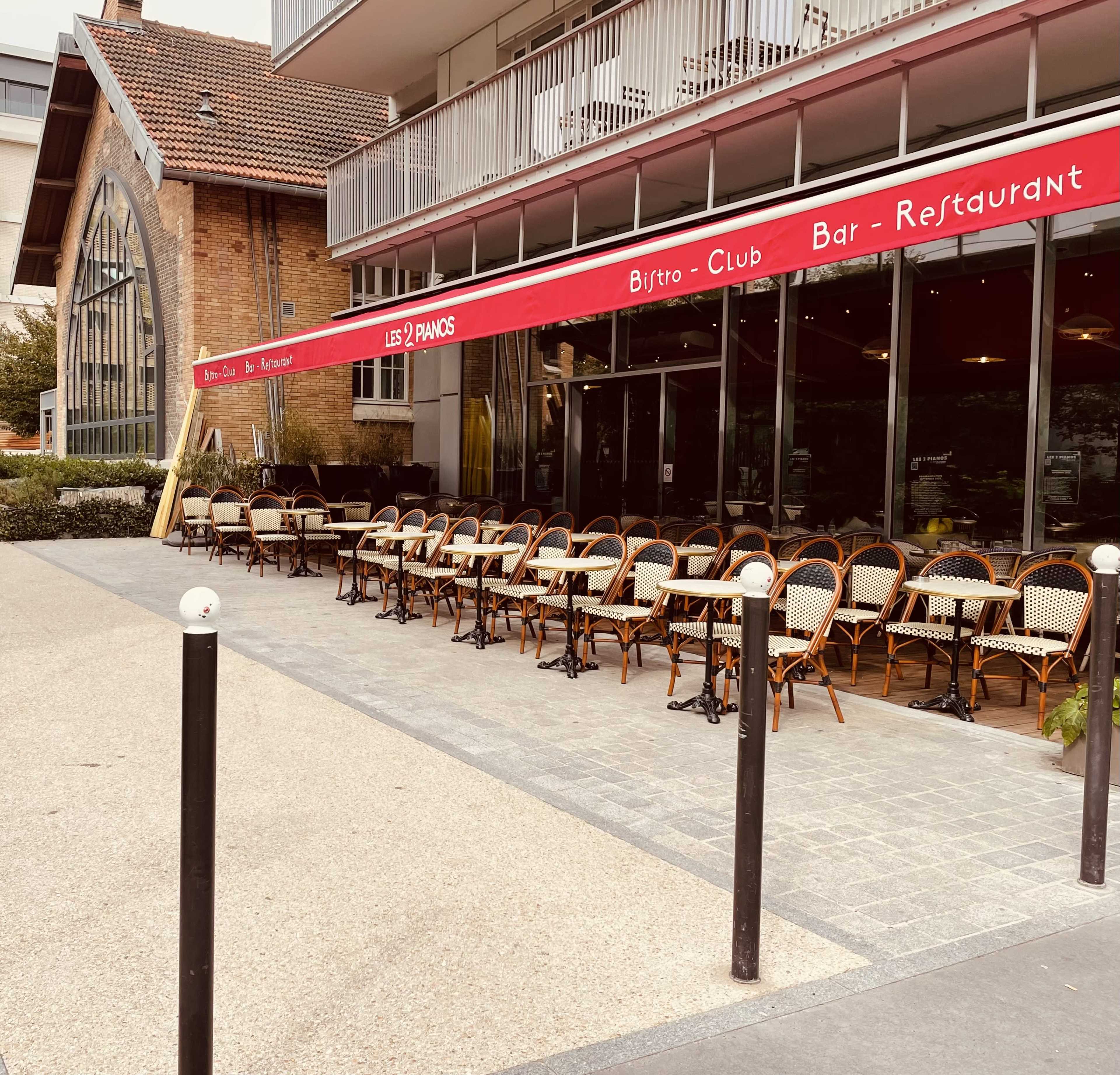 A restaurant with a red awning and rows of empty outdoor seating in front of a glass facade.