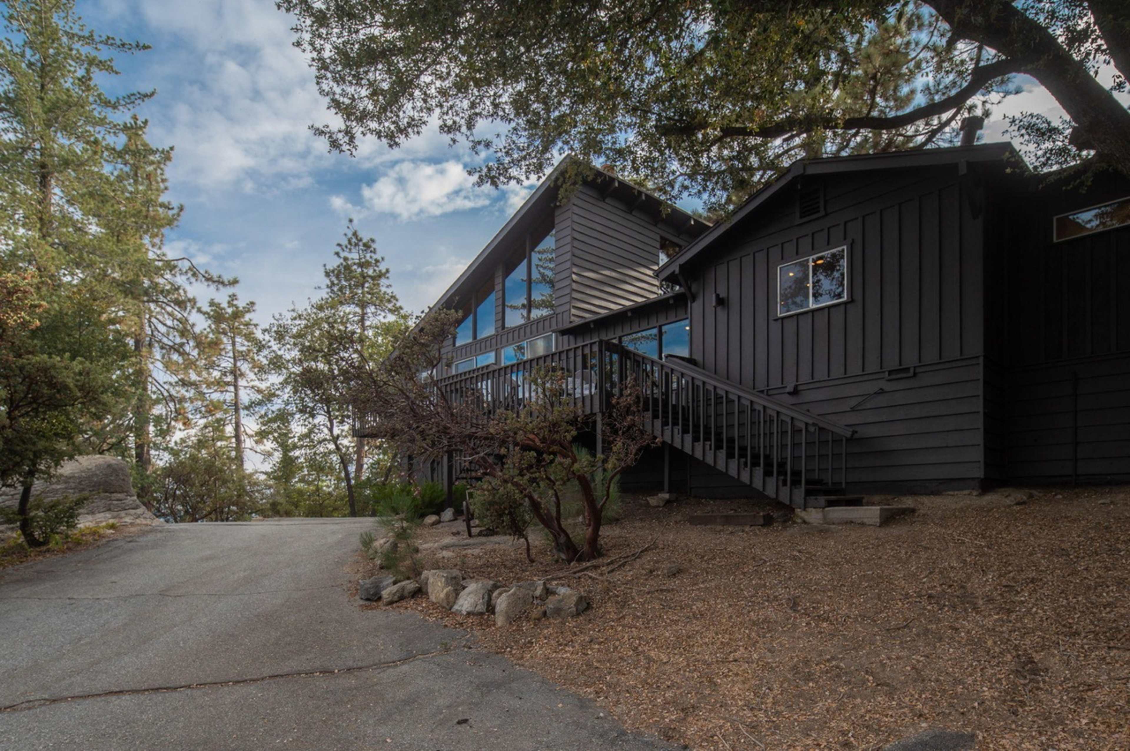 A dark wooden house is situated on a gravel pathway lined with trees and shrubs.