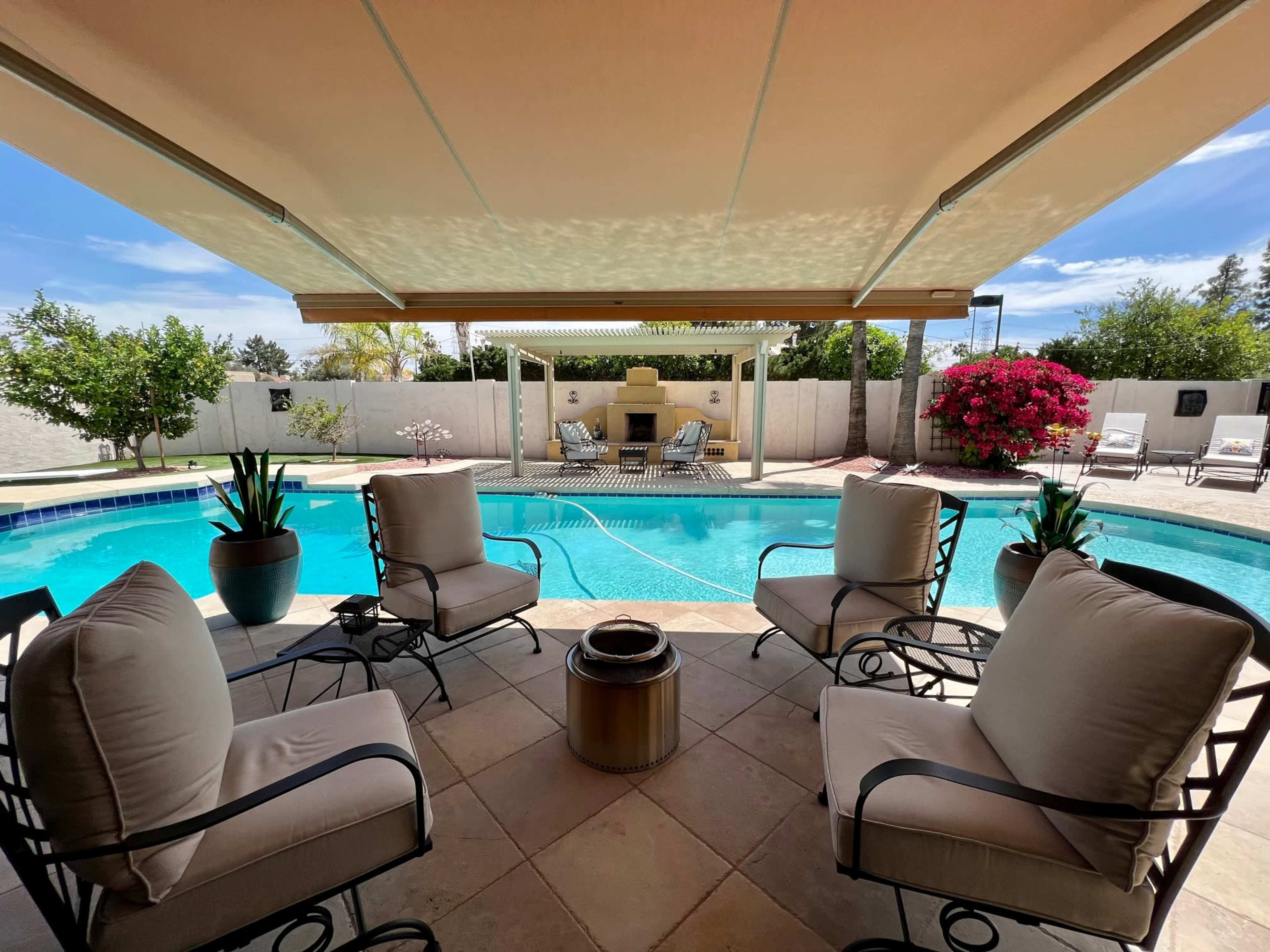 A shaded seating area with four chairs surrounds a swimming pool, framed by blooming plants and a patio enclosure.
