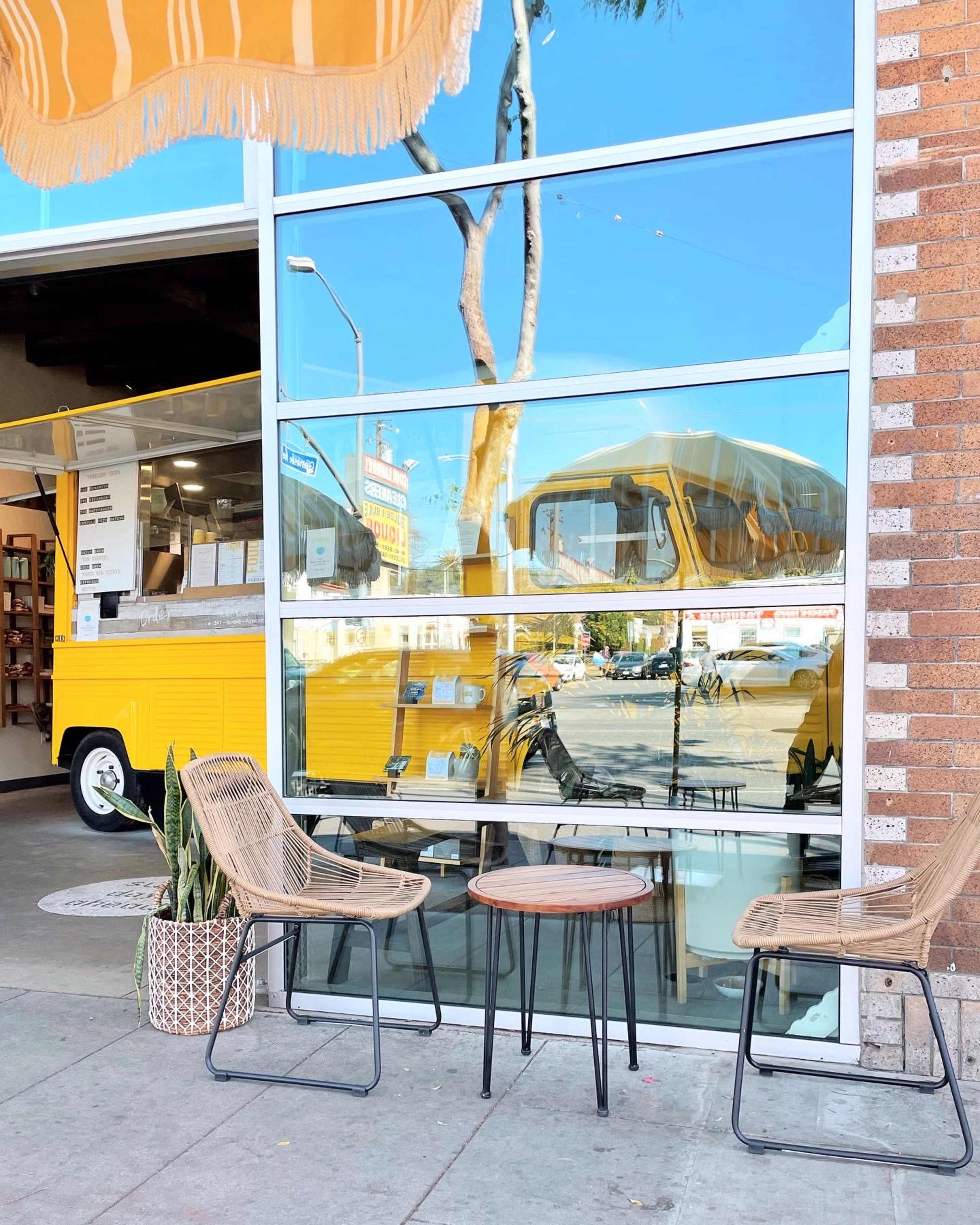 The image shows a storefront with large glass windows reflecting a yellow food truck and two chairs with a small table outside.