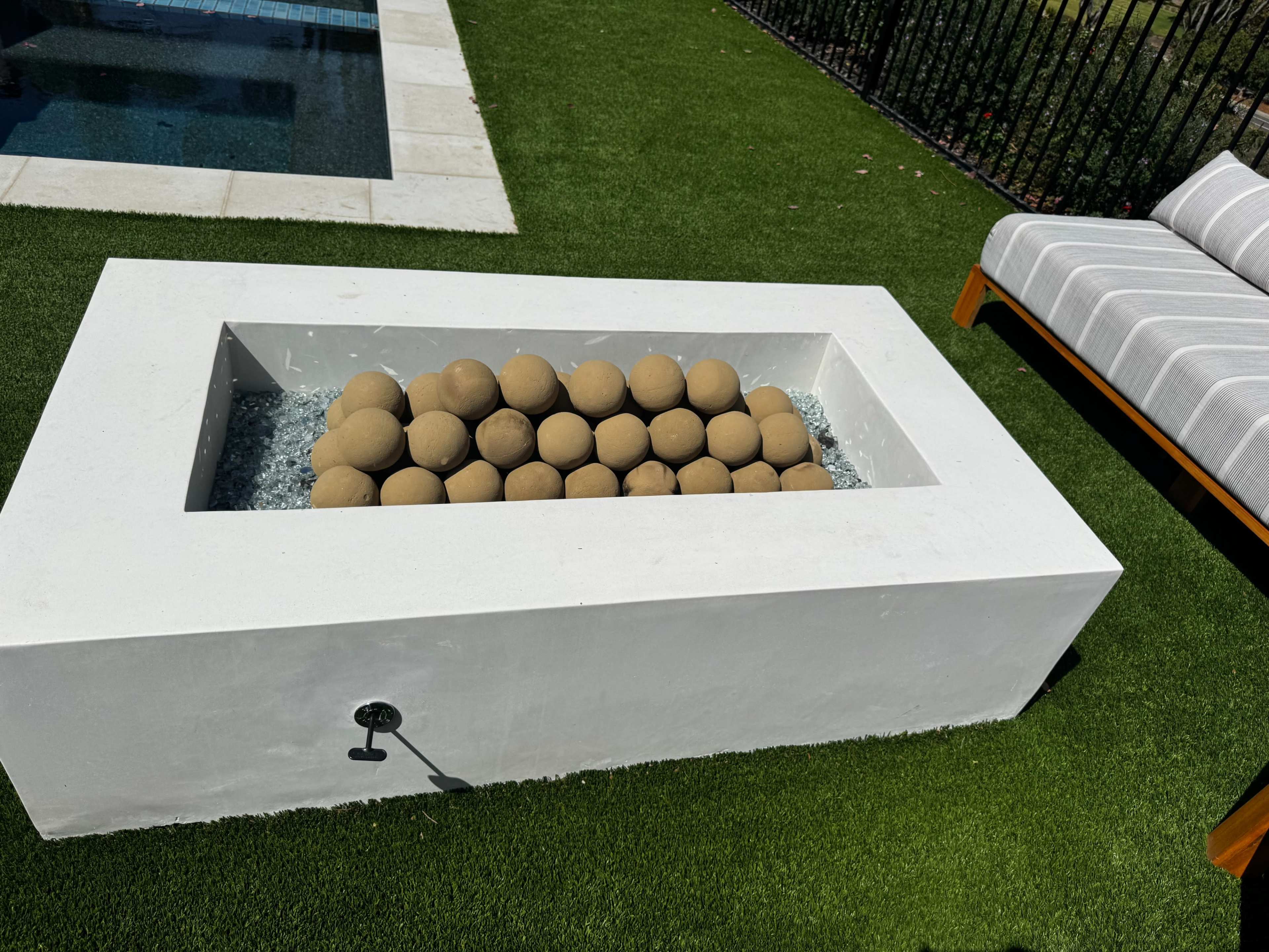 The image shows a rectangular outdoor table containing rounded brown stones arranged in a bed of clear glass, situated on green artificial grass near a swimming pool and a lounge chair.