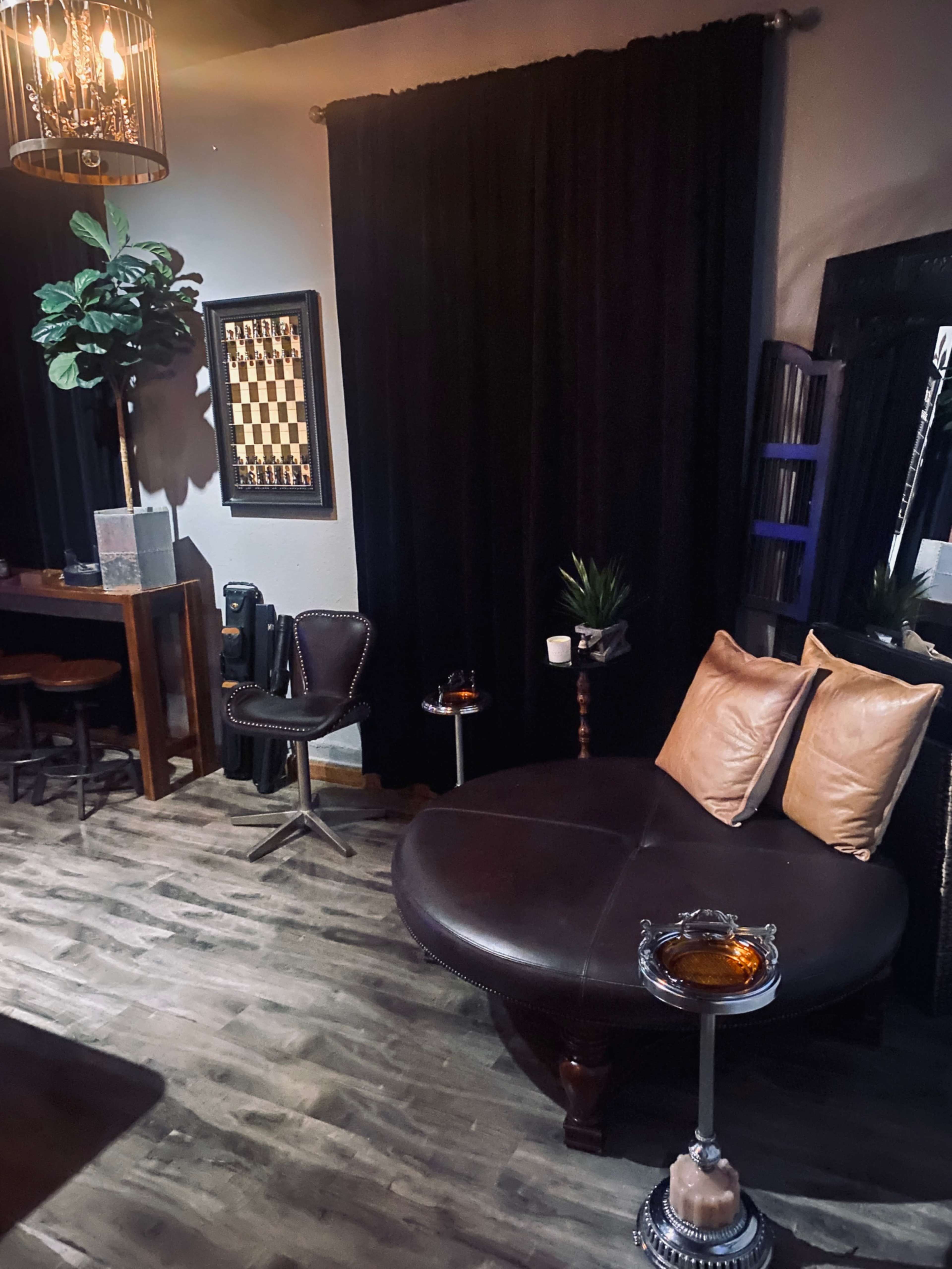 The image shows a cozy lounge area featuring a curved leather sofa with two pillows, a small side table, a plant, and a dark backdrop, illuminated by a chandelier.