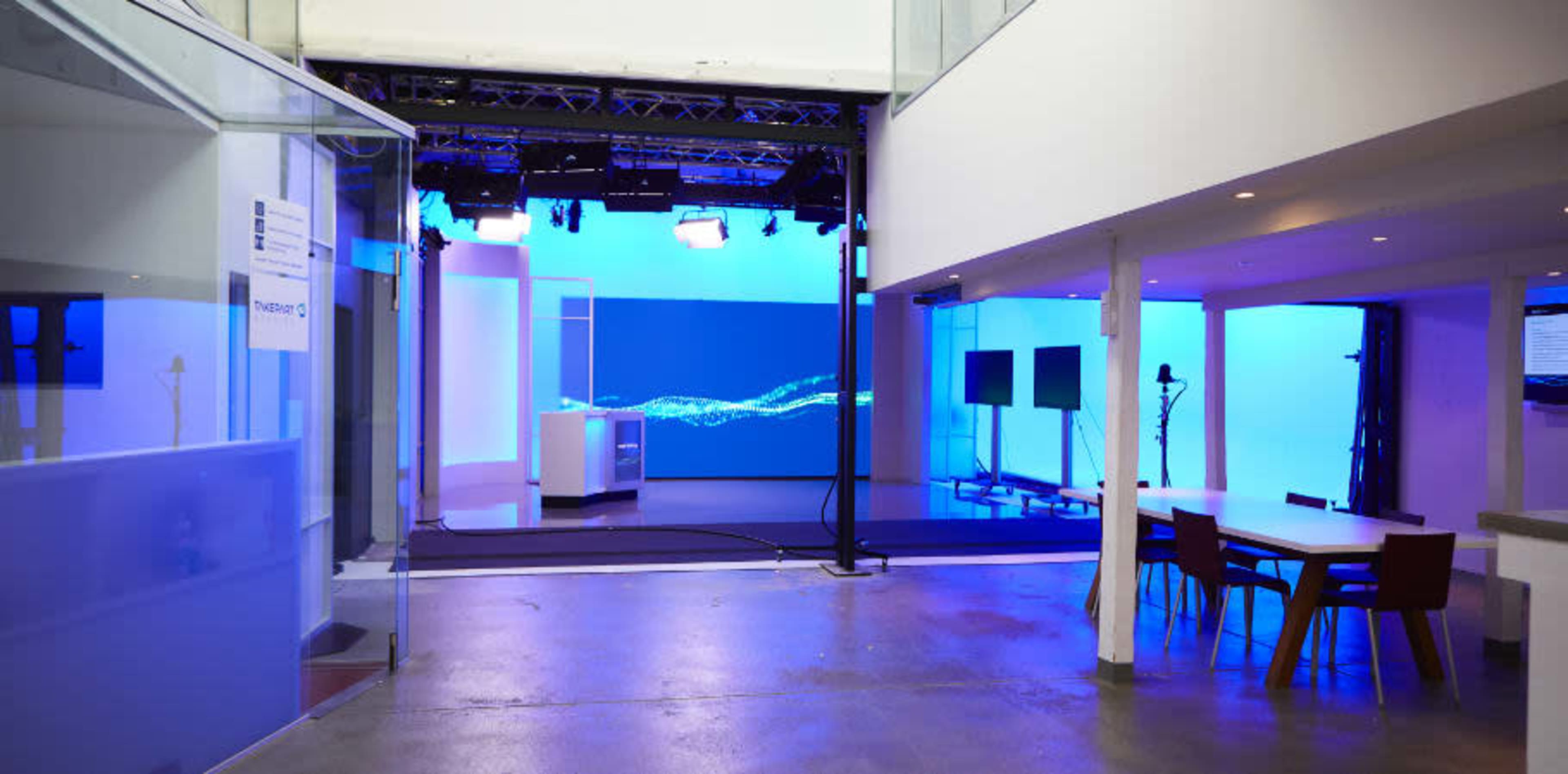 The image shows a modern studio space with blue lighting, featuring a presentation area, screens, and a dining table with chairs.