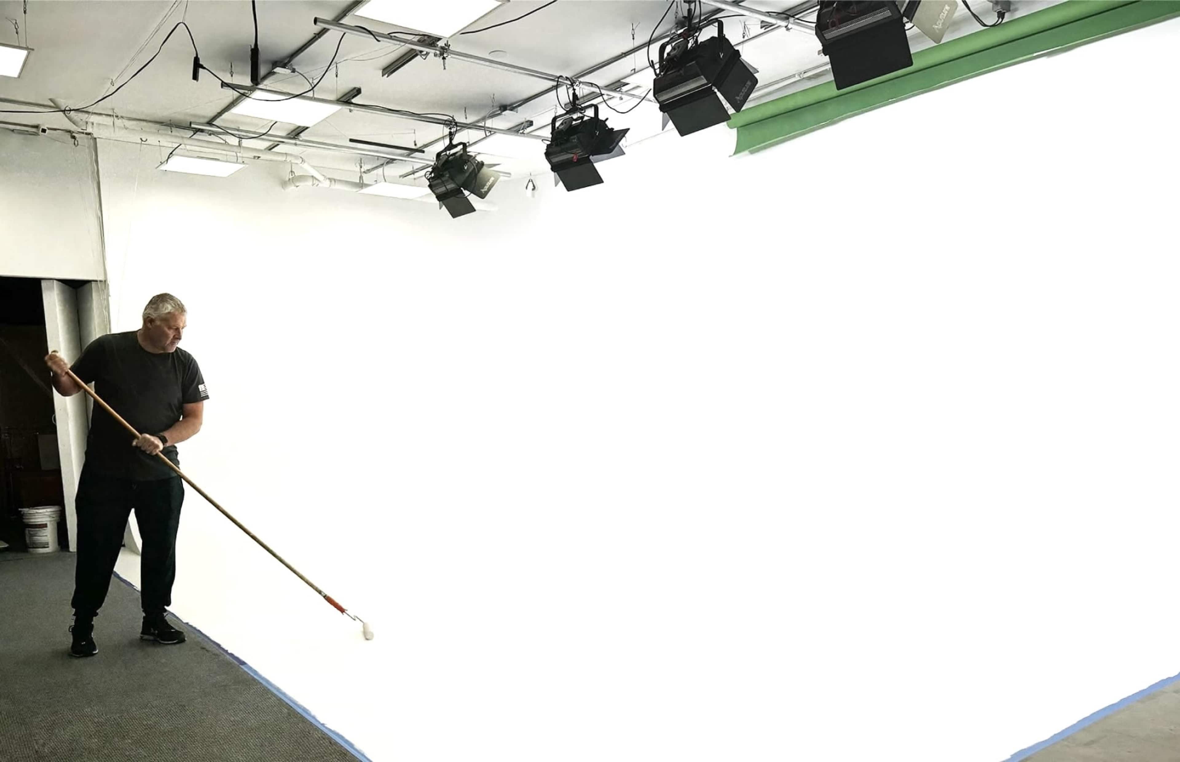 A person is painting a large, white wall in a studio space that is lit by overhead lights.