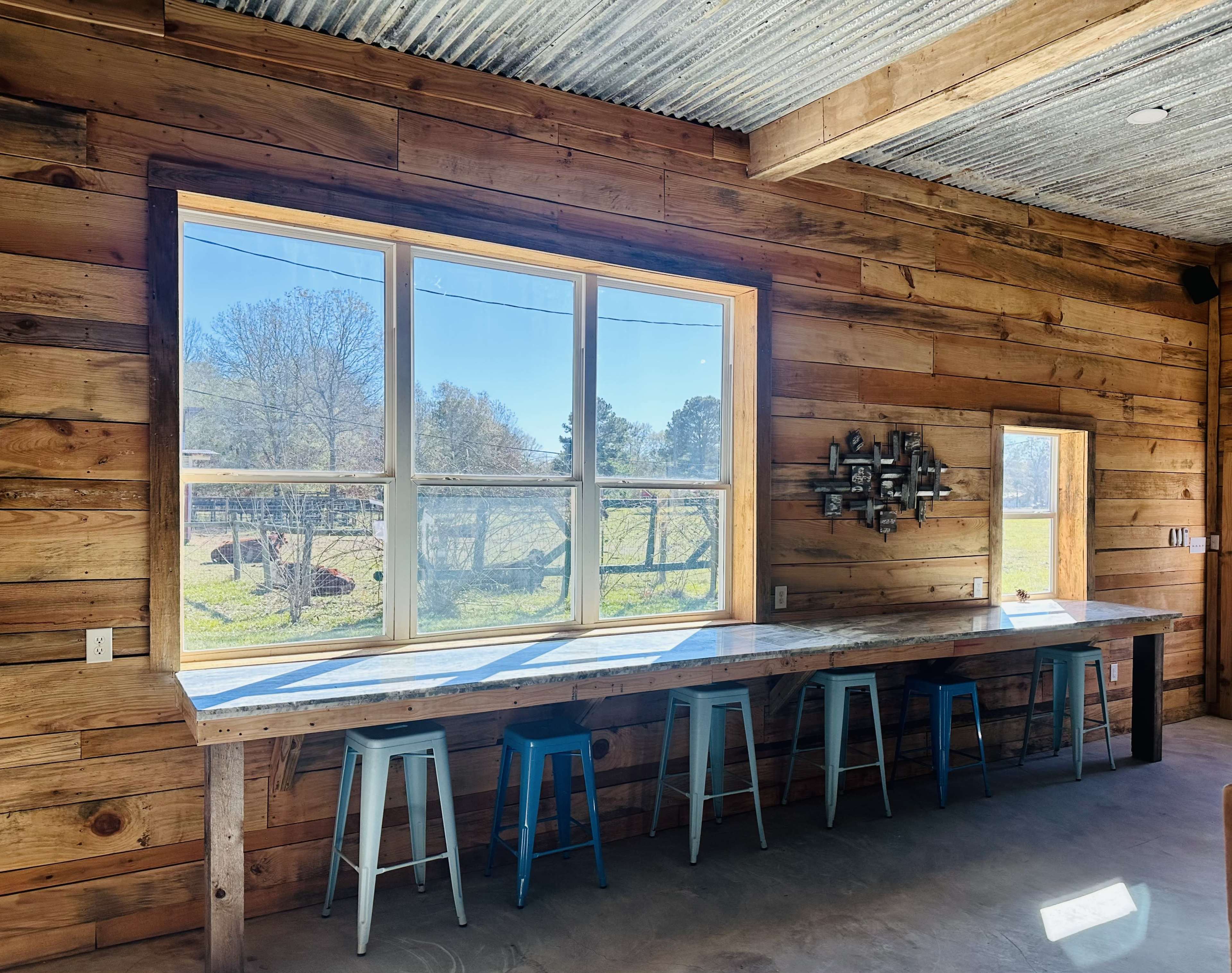 A large window frames a view of trees outside, illuminating a wooden interior with a table and blue stools.
