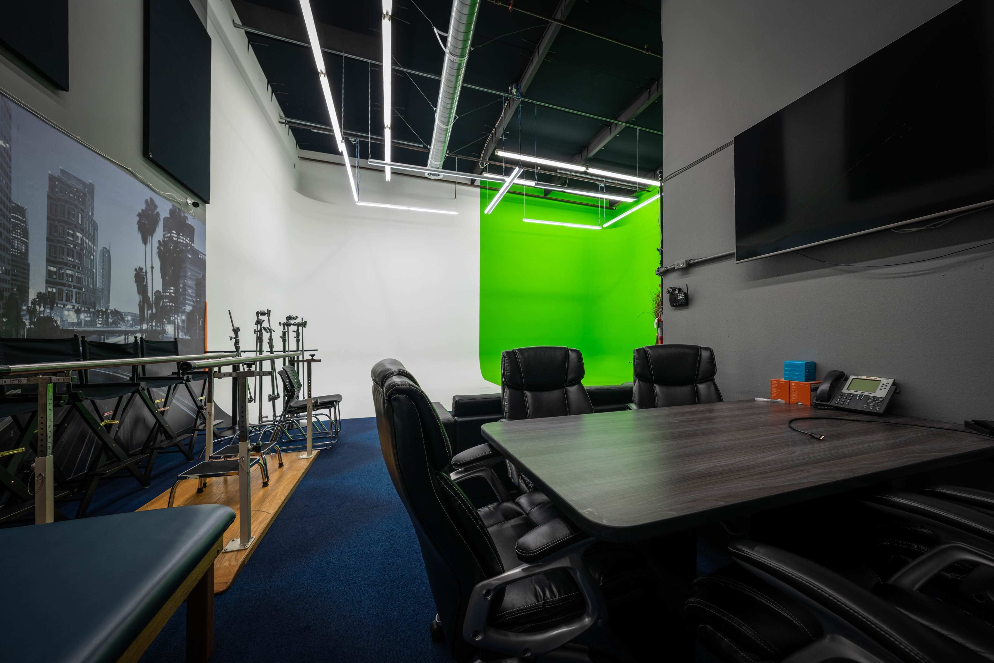A meeting room features a dark wooden table surrounded by black leather chairs, with a green screen on the wall and a large monitor mounted nearby.