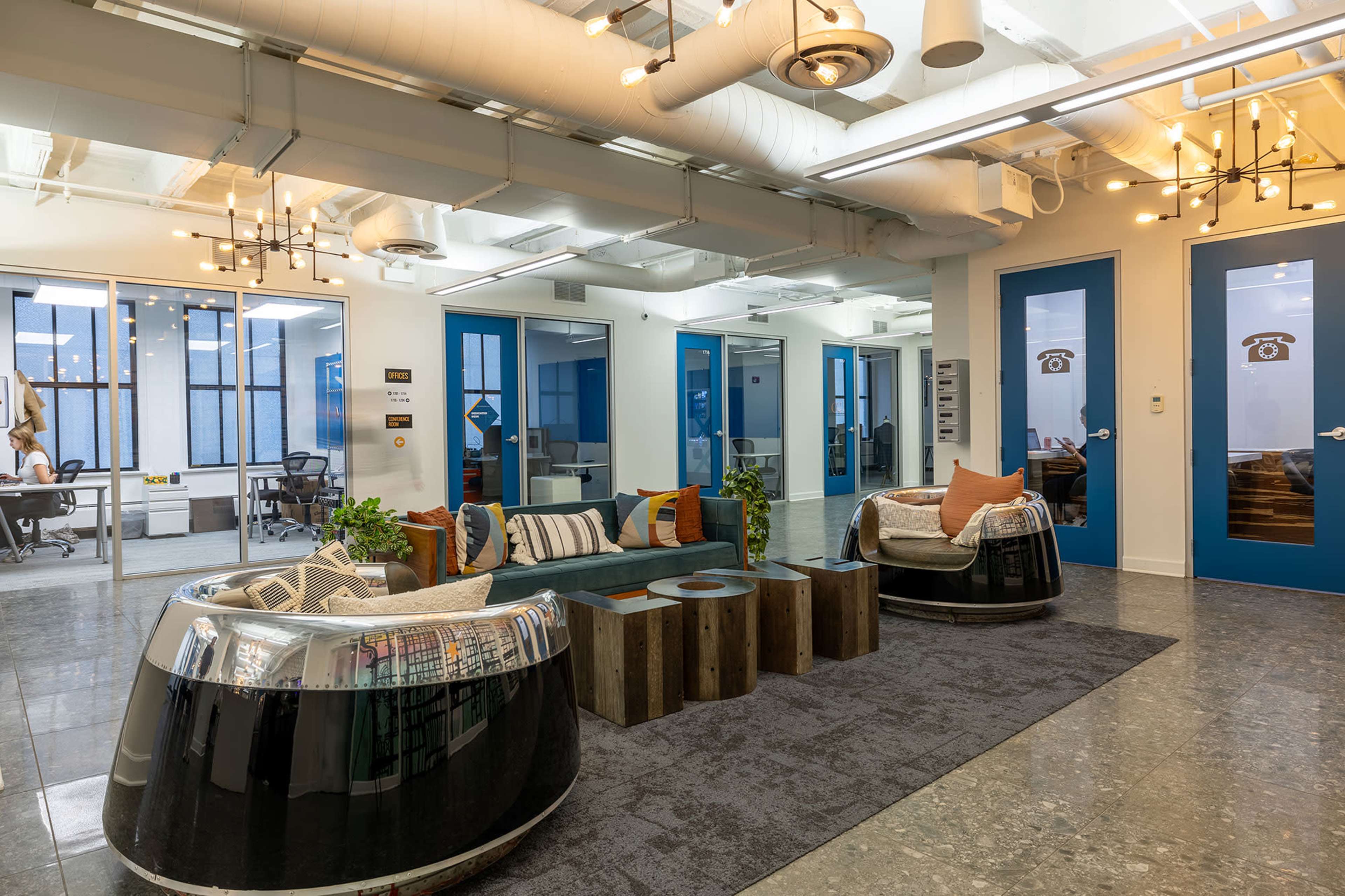 The image shows a modern office space featuring a cozy seating area with two unique chairs, a low table made of wooden crates, and blue doors leading to other workspaces.