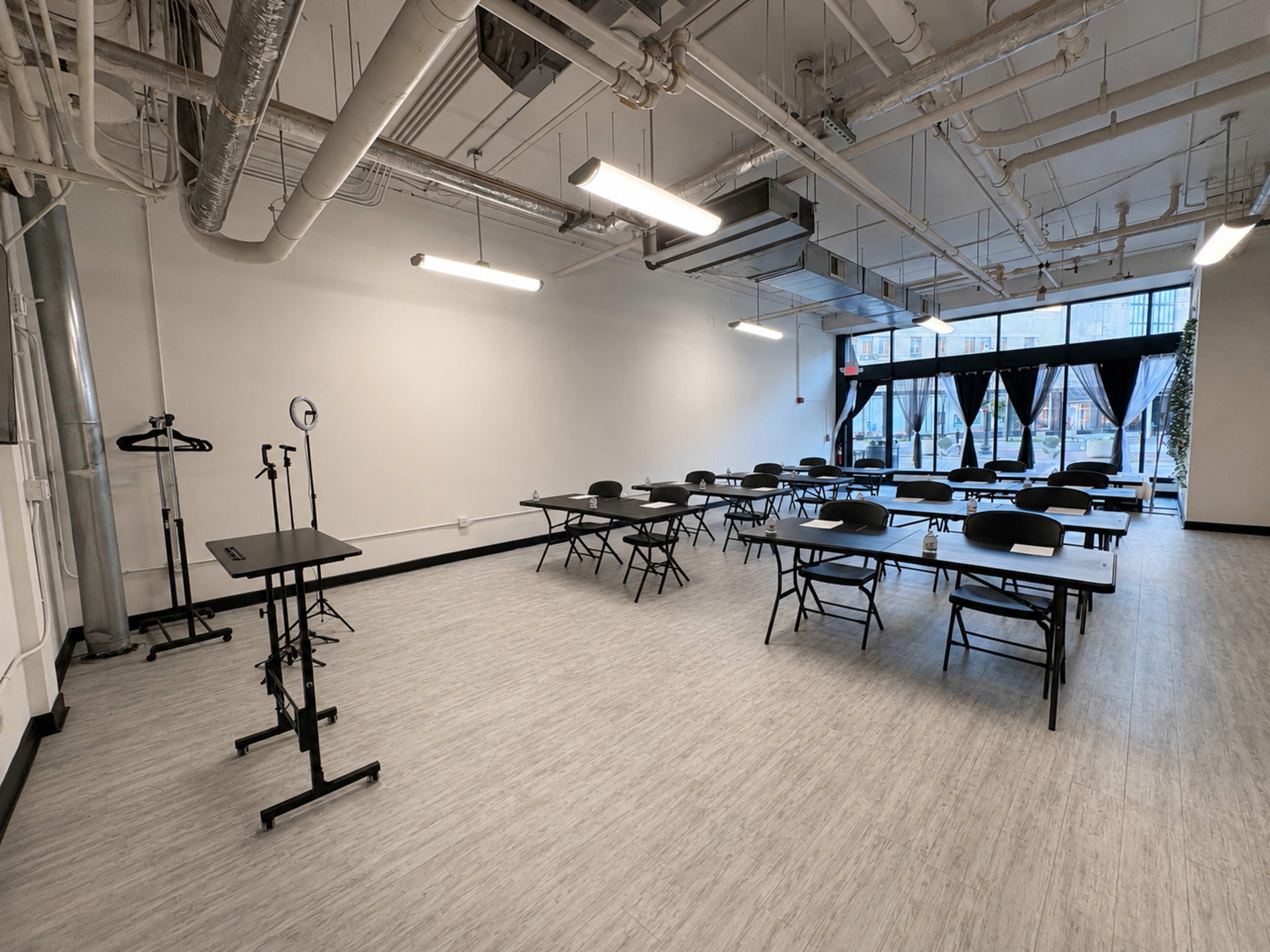 The image shows a spacious, minimalistic room with several tables and chairs arranged for a meeting or event, with large windows along one wall.