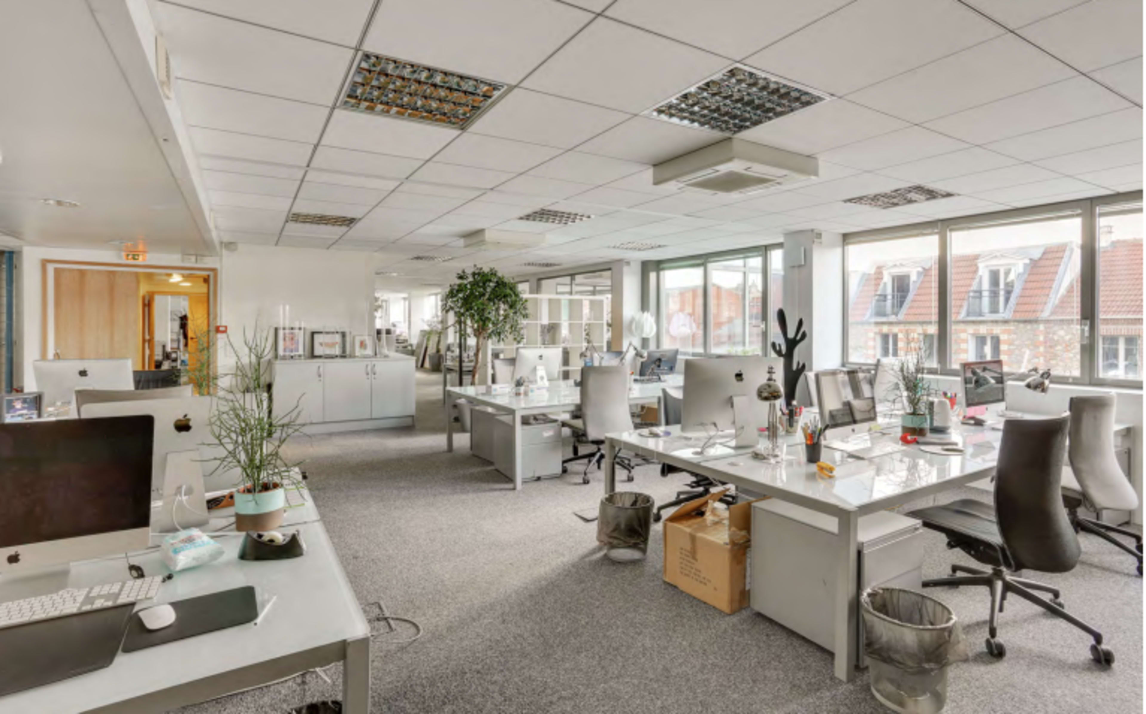 The image shows a modern office space with multiple desks, computer stations, and potted plants, illuminated by natural light from large windows.