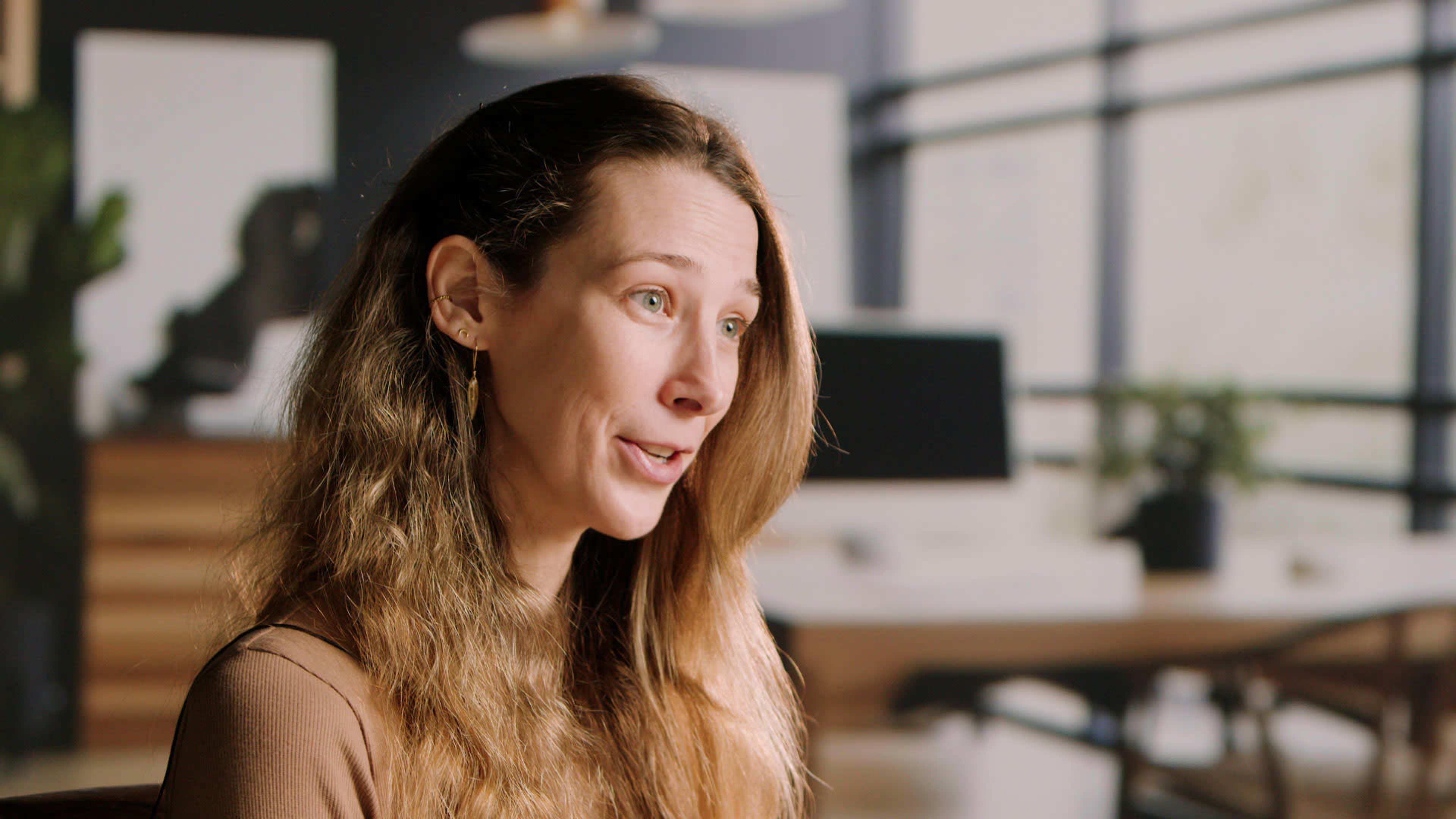 A woman with long, wavy hair is speaking in an office setting with furniture and greenery in the background.