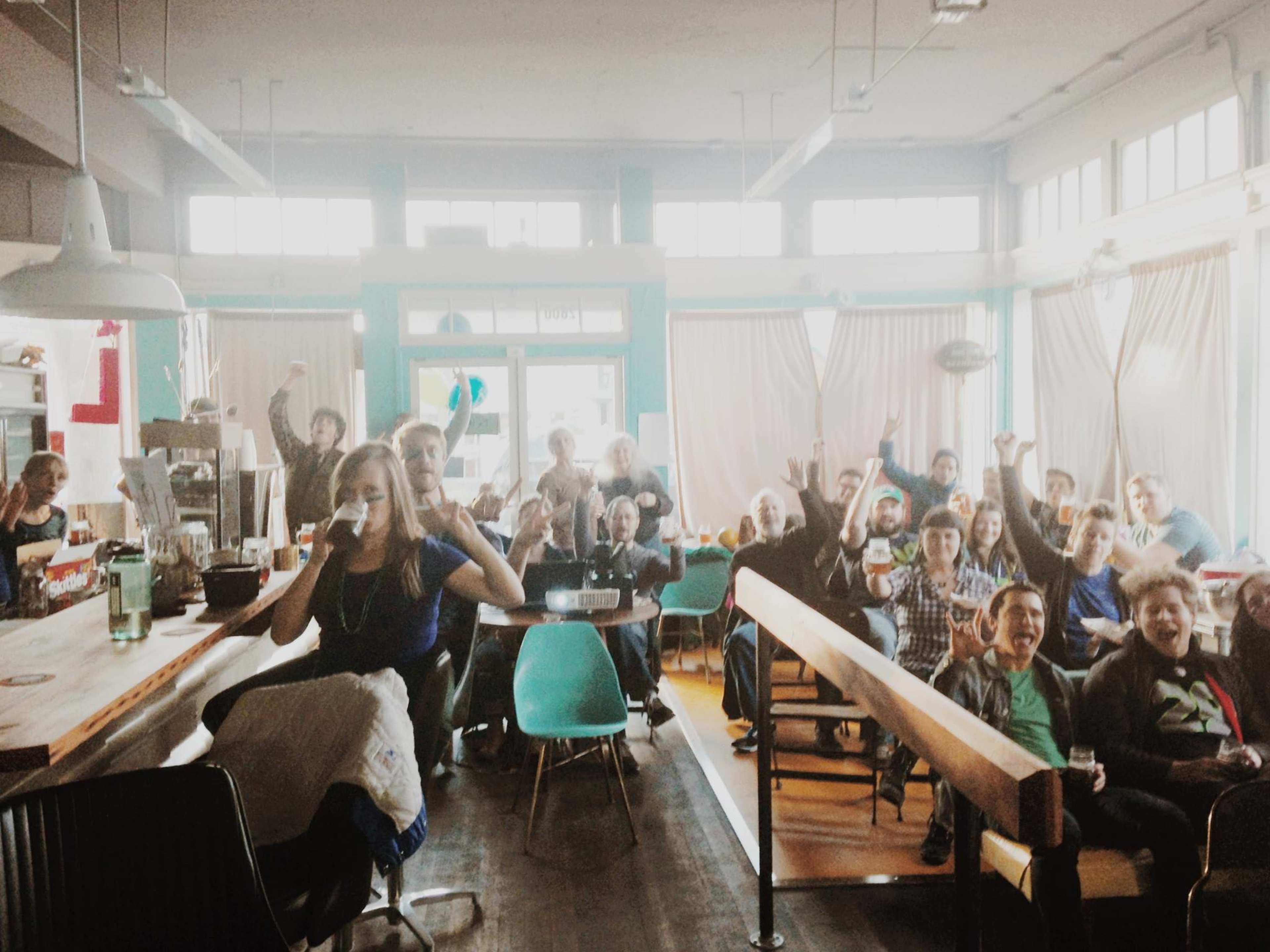 A large group of people is gathered in a lively cafe, smiling and cheering towards the camera.