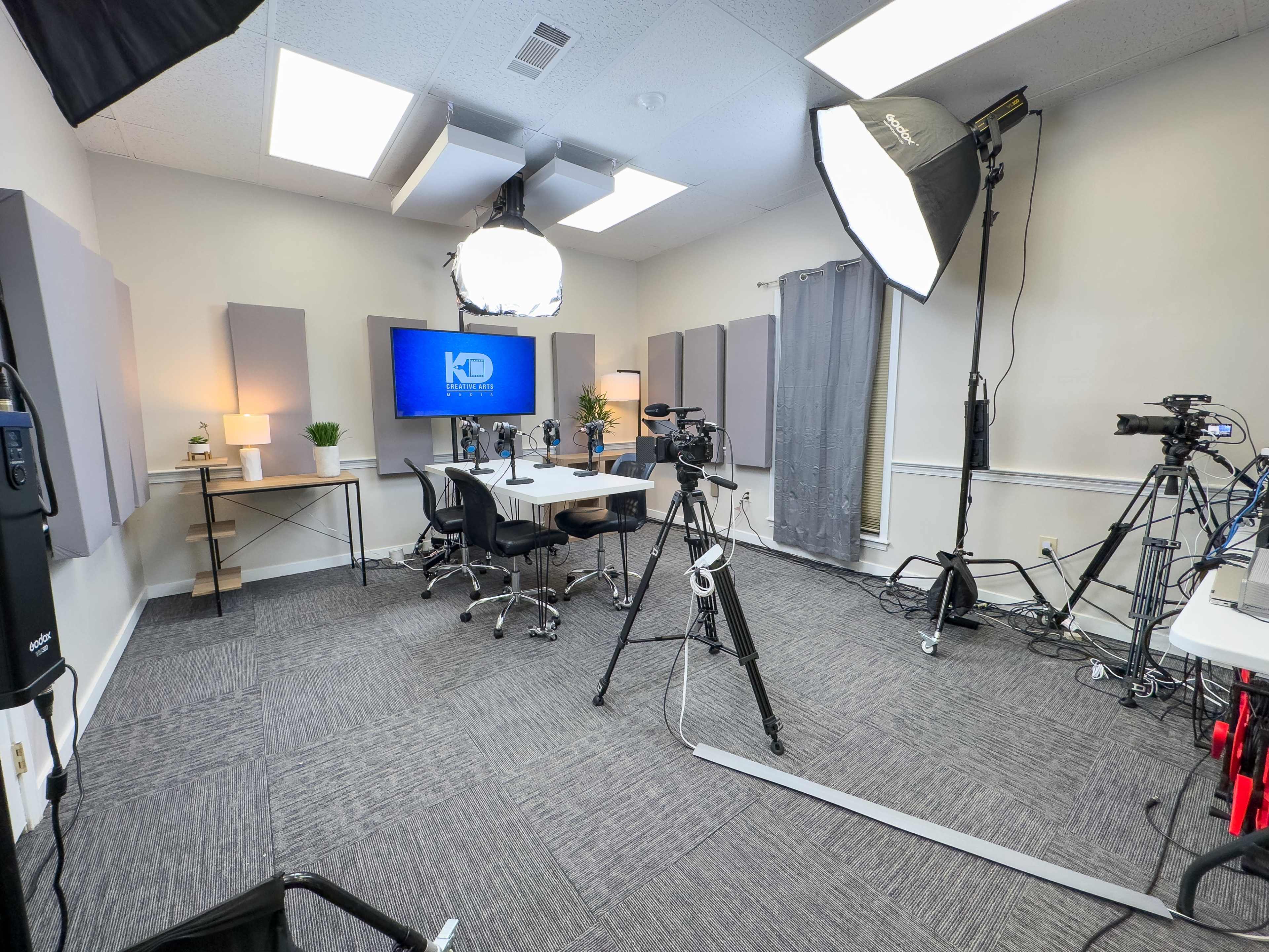 A well-equipped video production studio with several cameras, lighting equipment, and a monitor set up around a central table and chairs.