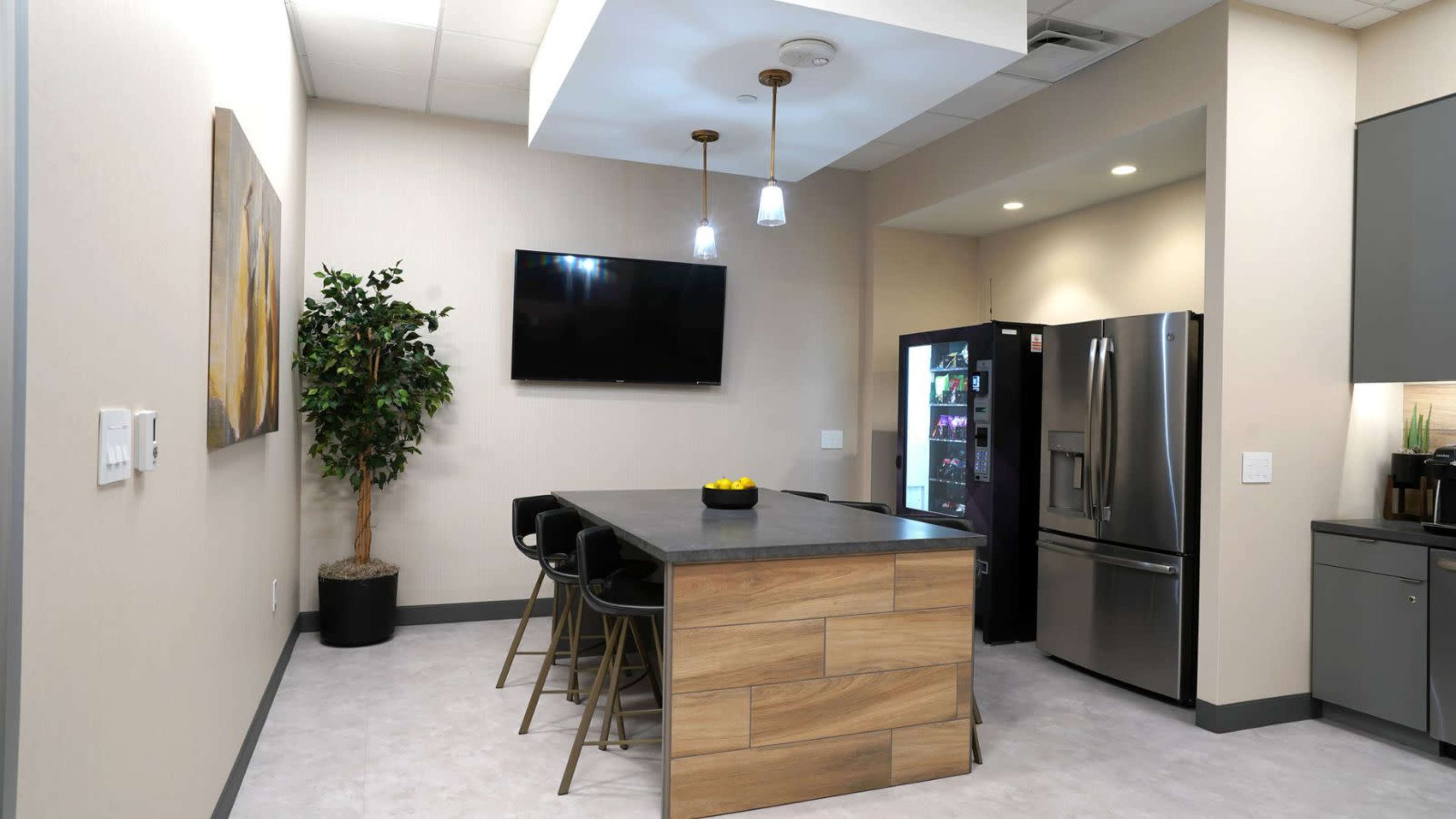 The image shows a modern kitchen with a central island, black bar stools, a television mounted on the wall, and a stainless steel refrigerator.