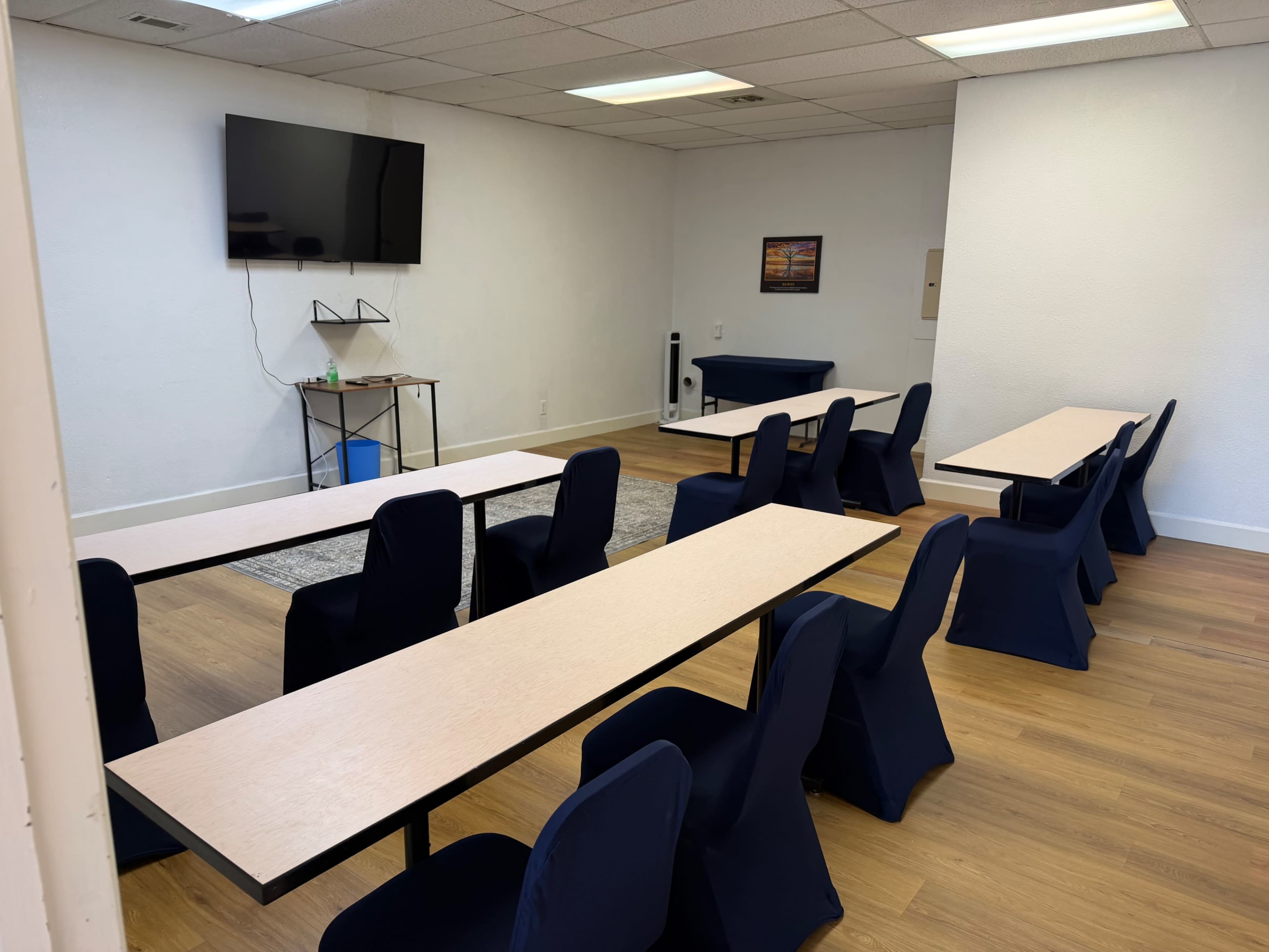 The image shows a classroom setting with several long tables and chairs arranged in a row, along with a television mounted on the wall.