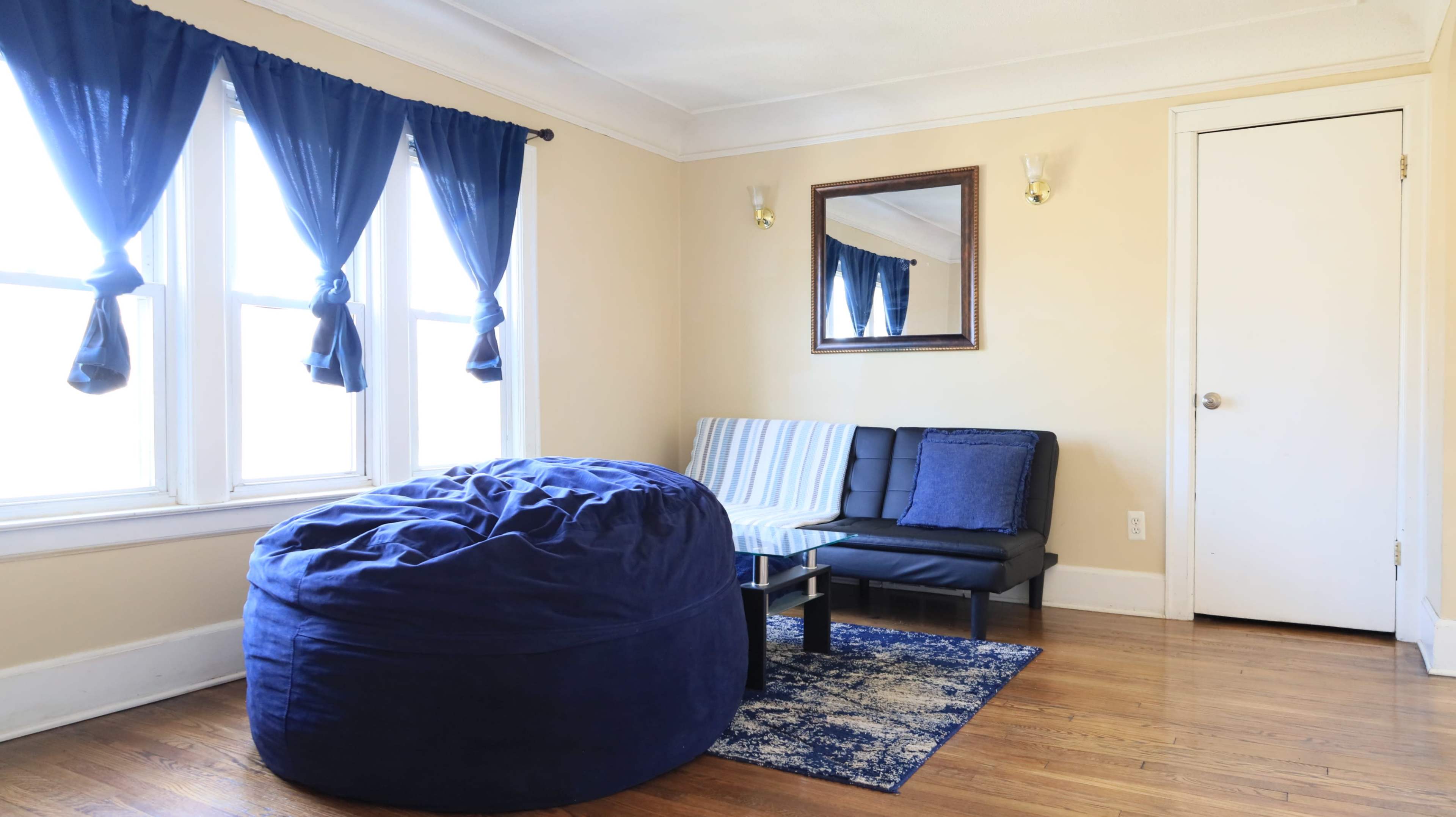 The image features a cozy living room with two windows dressed in blue curtains, a large blue bean bag chair, a black folding sofa, and a mirror on the wall.