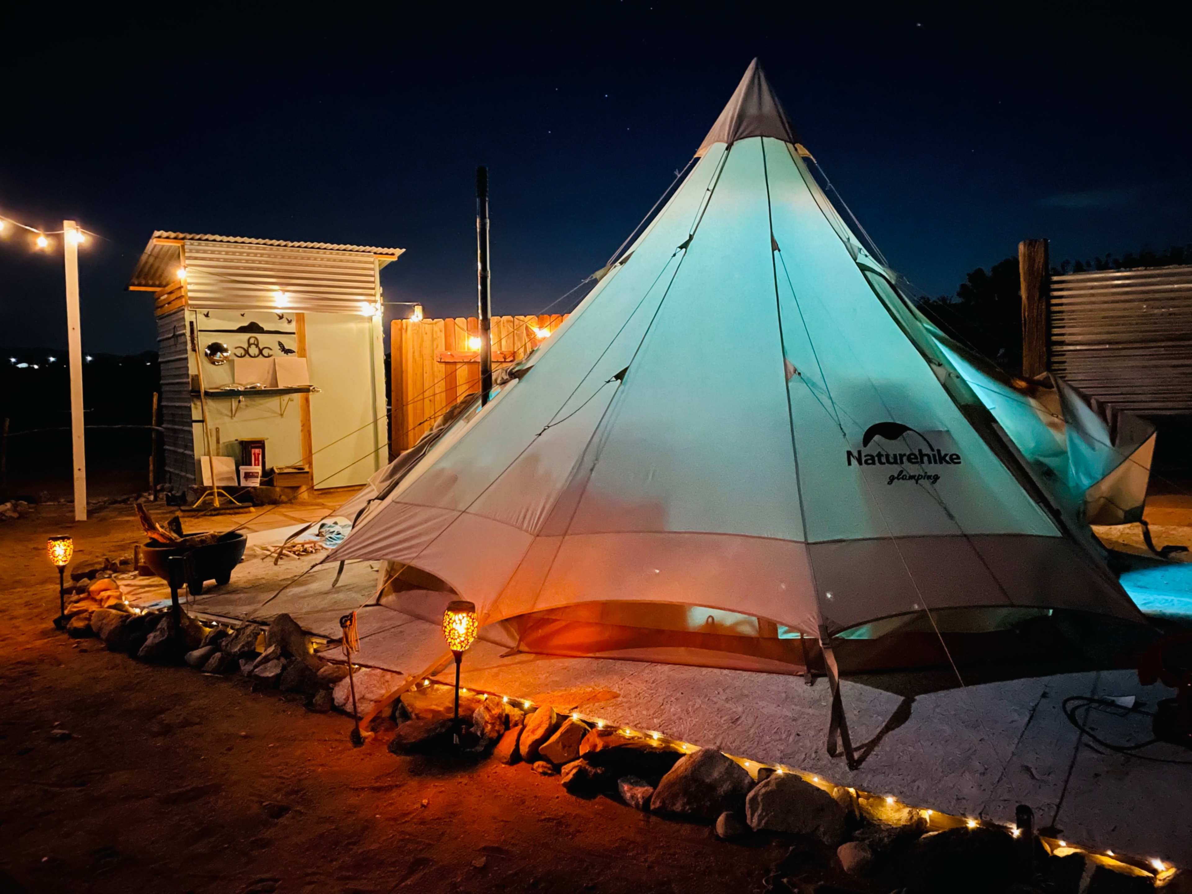 A large, illuminated camping tent sits in a desert area at night, surrounded by decorative lights and wooden structures.
