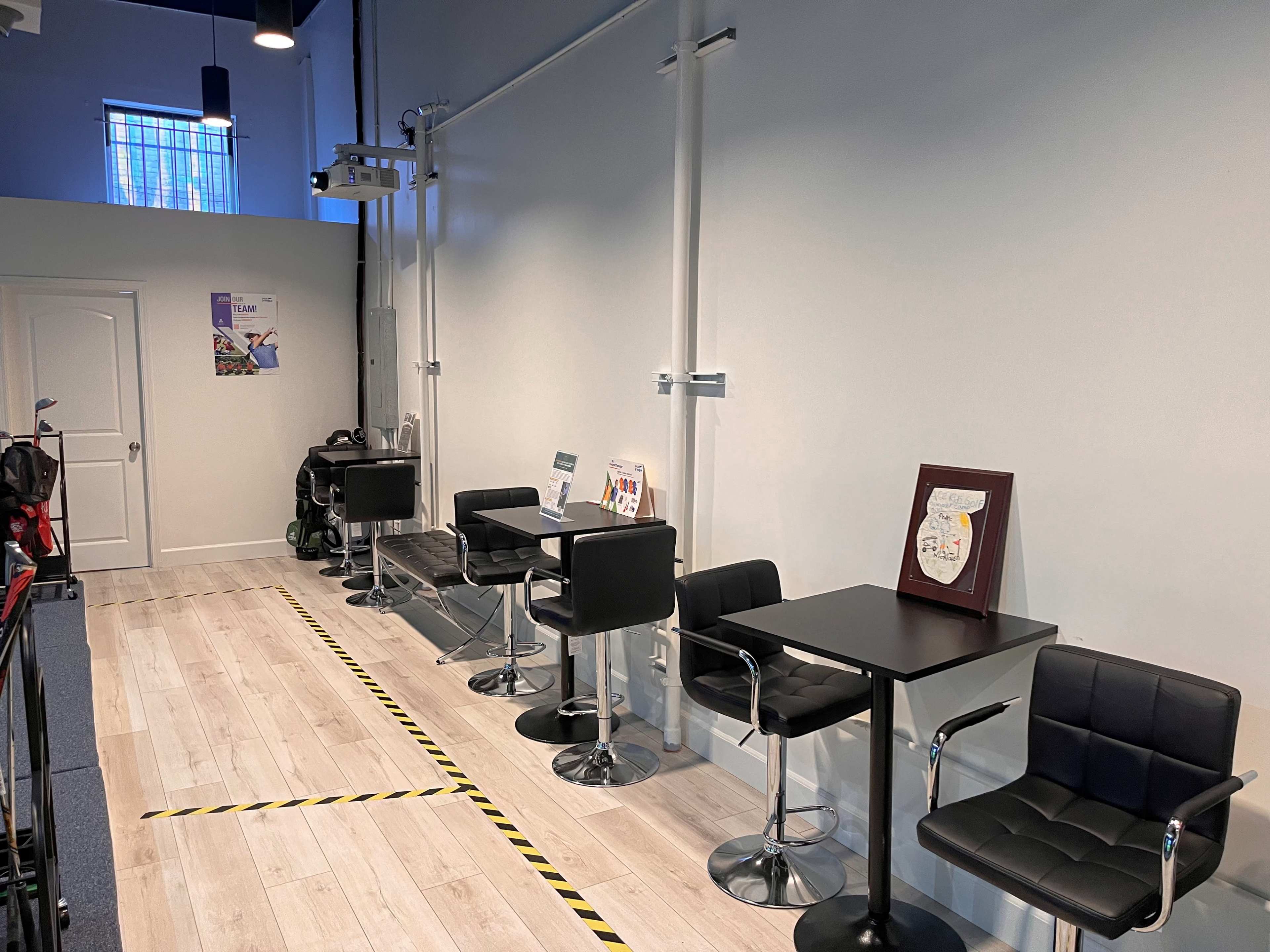 A well-lit interior space with a row of black chairs and tables along a white wall, separated by yellow caution tape on the floor.
