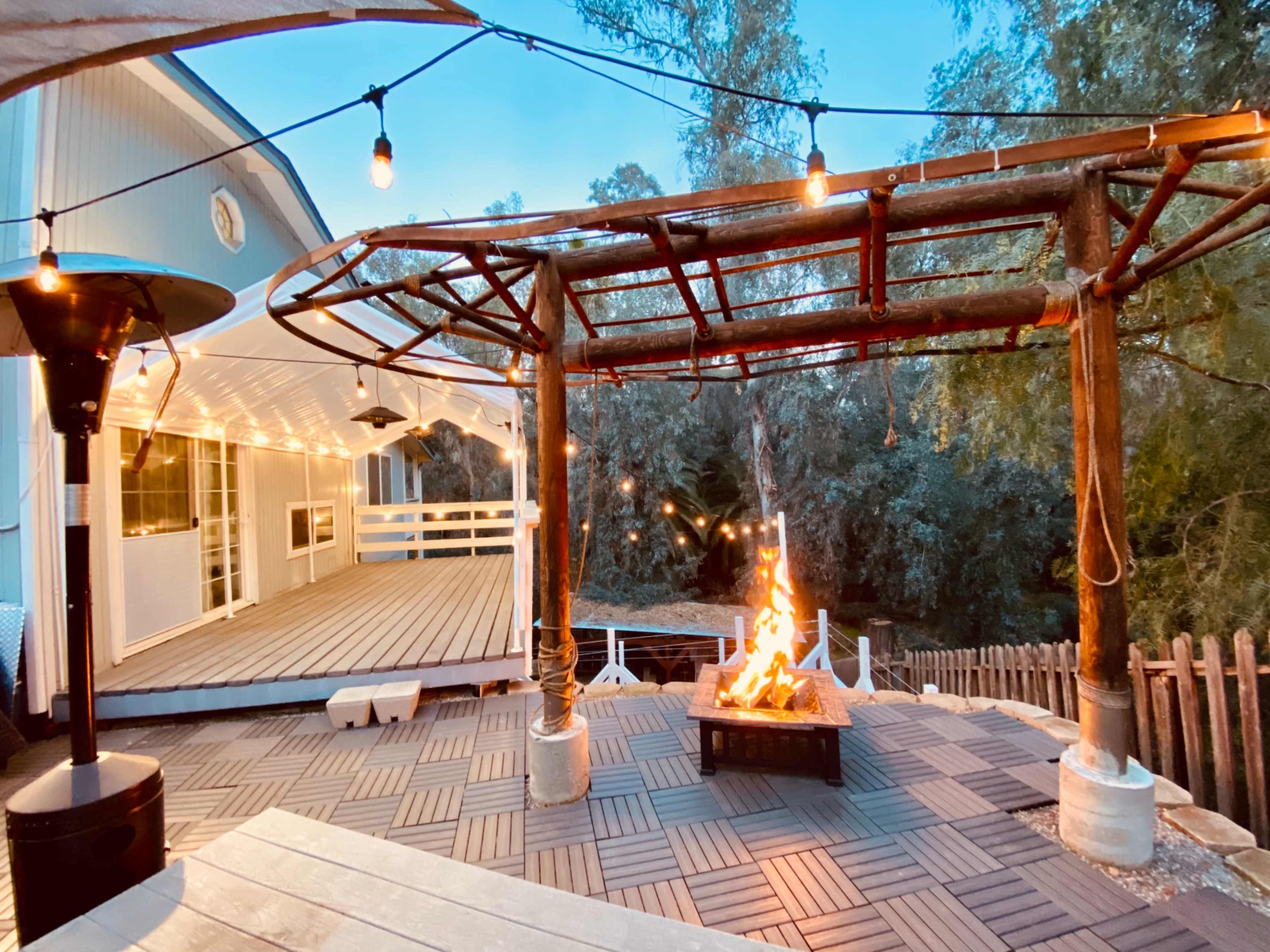 A wooden pergola with string lights surrounds a fire pit on a patio beside a house.