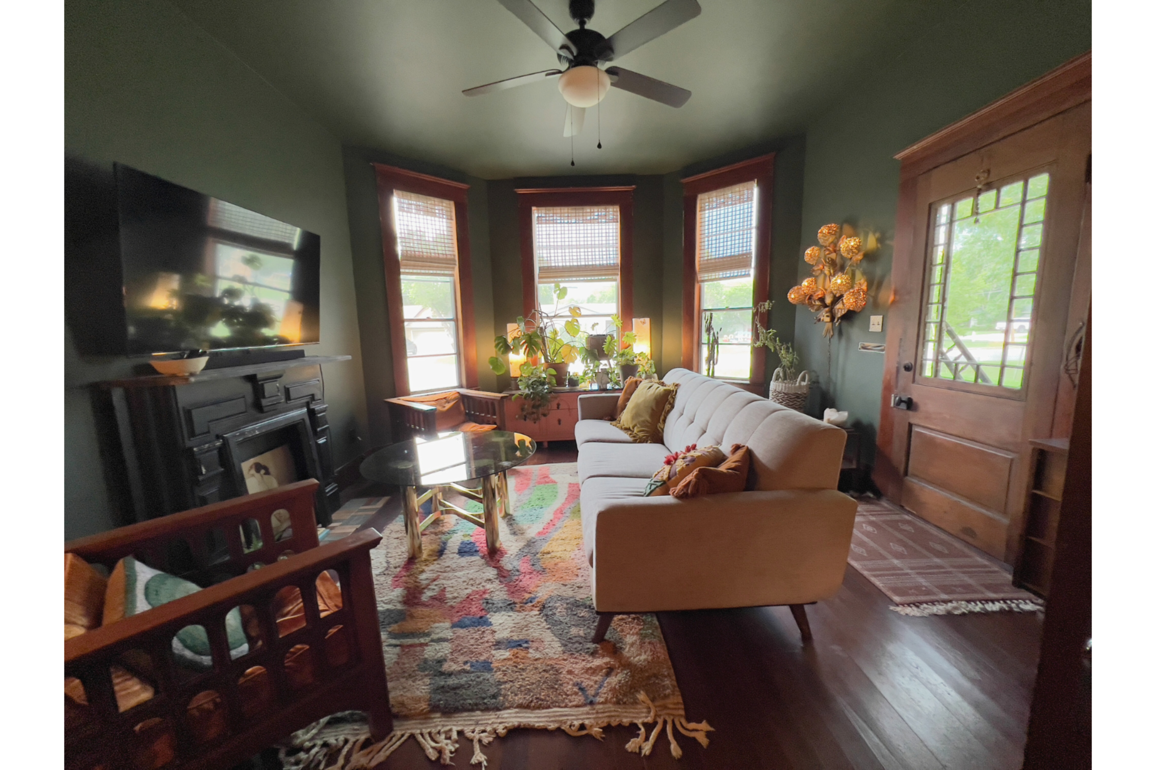 The image shows a cozy living room with a sectional sofa, a coffee table, a decorative rug, and several windows allowing natural light to illuminate the space.