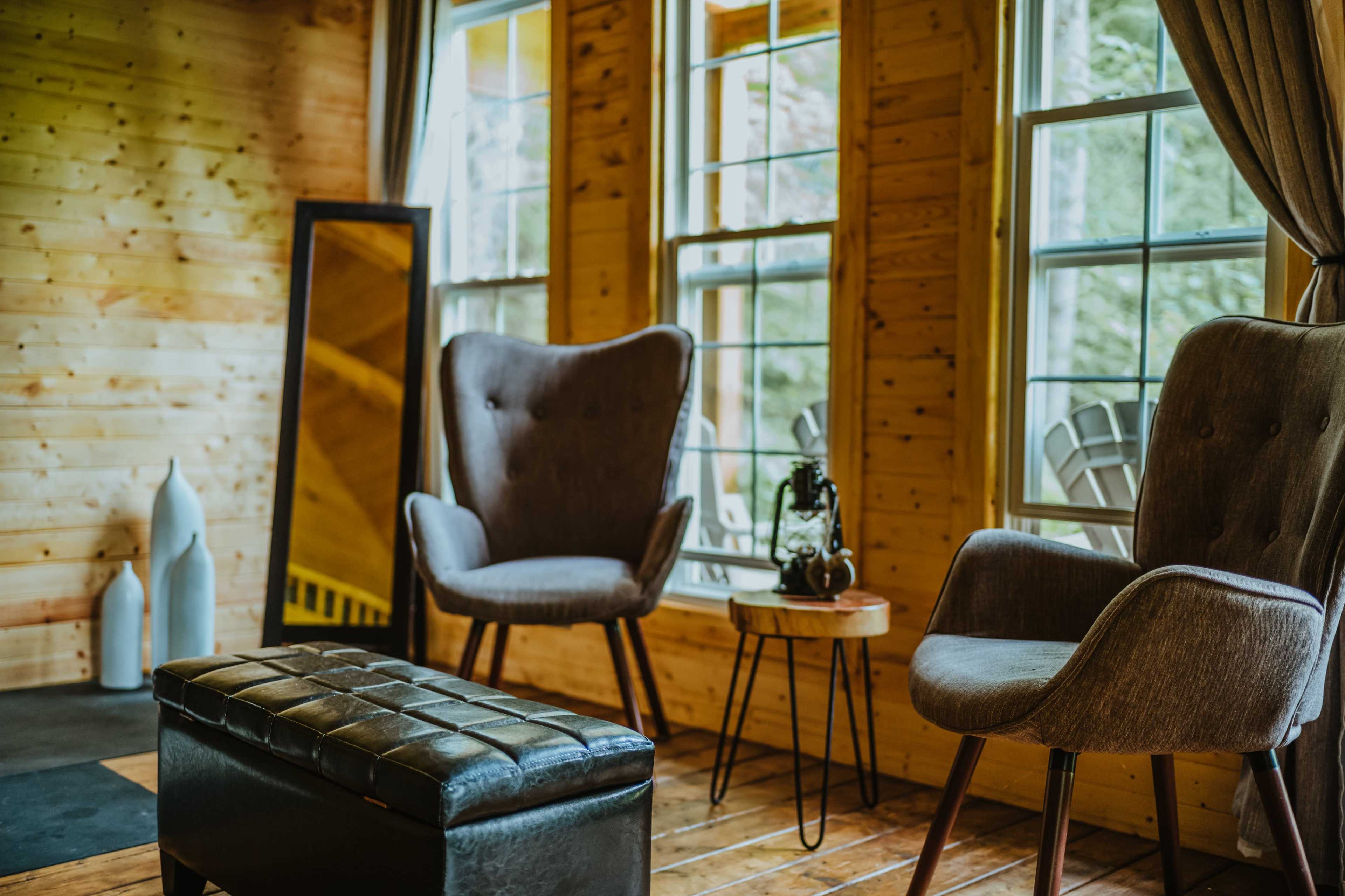 The image shows a cozy interior with two armchairs, a small round table, a mirror, and large windows allowing natural light to enter.