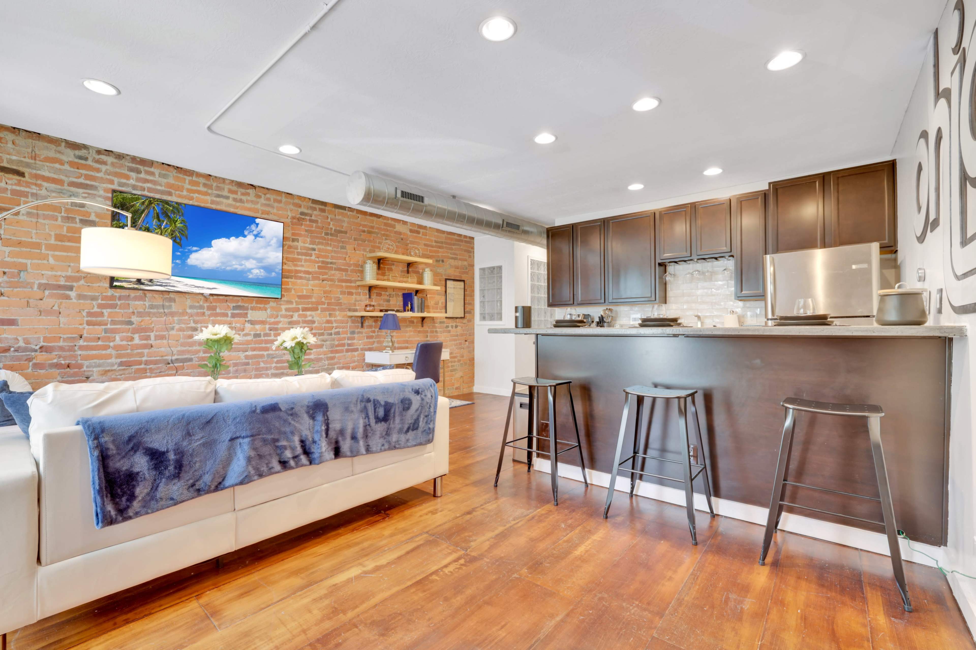 A modern living space with a brick wall, a white sofa adorned with a blue throw, a kitchen area featuring dark cabinetry, and bar stools at the counter.