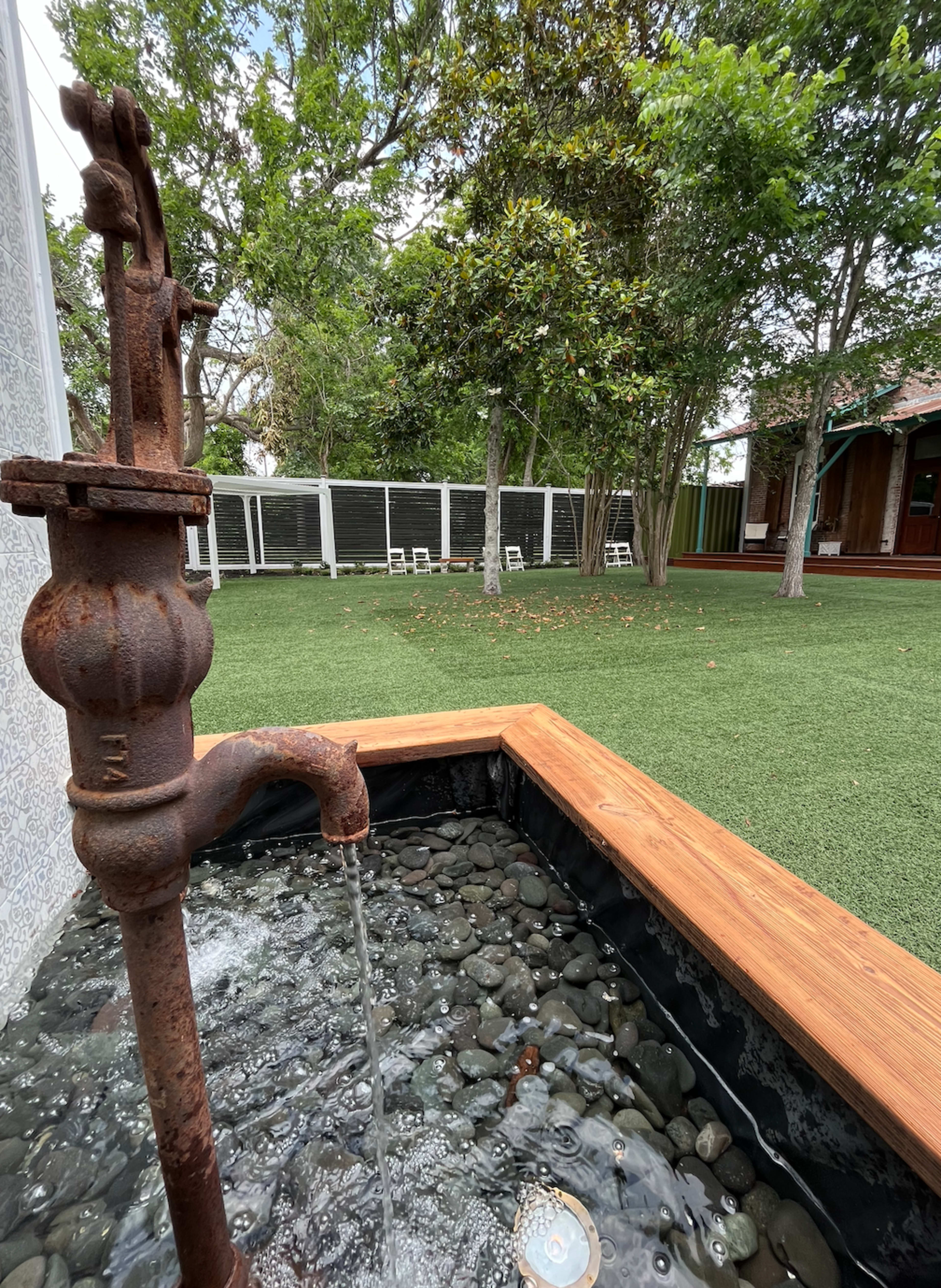 A rusty water pump sits beside a stone-lined basin, with water flowing into it amidst a green lawn and trees in the background.