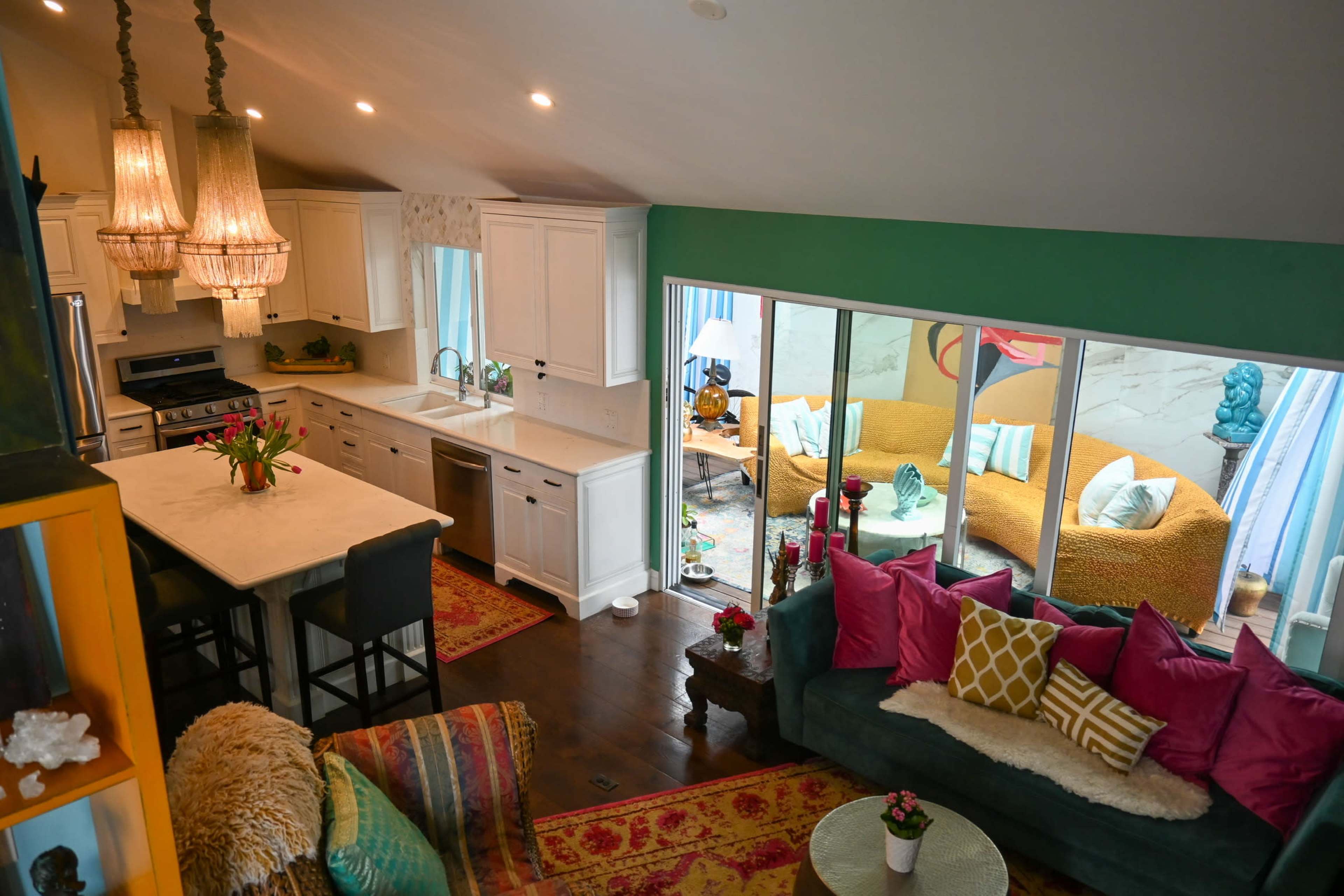 A bright, open-concept living space featuring a kitchen with white cabinetry, a cozy sitting area with a teal sofa, and a patio visible through large sliding glass doors.