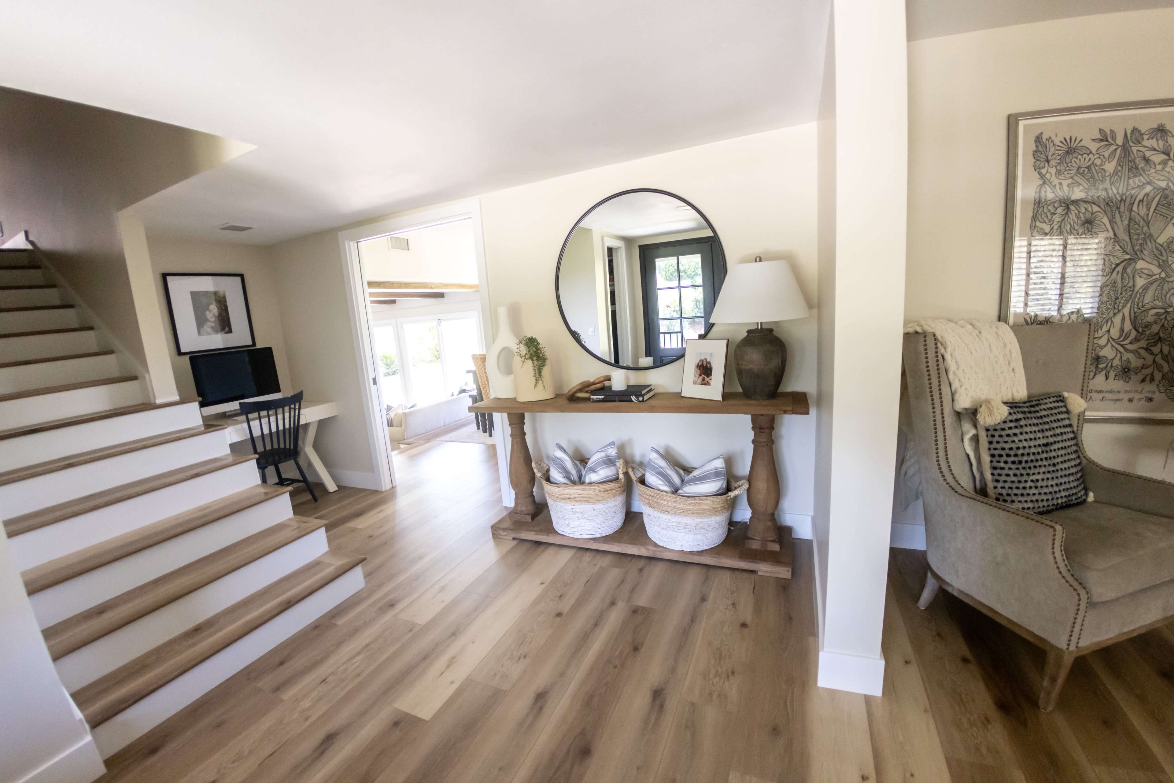 A bright, open interior features a staircase on the left, a round mirror above a wooden console table with decorative baskets, and a cozy seating area on the right.
