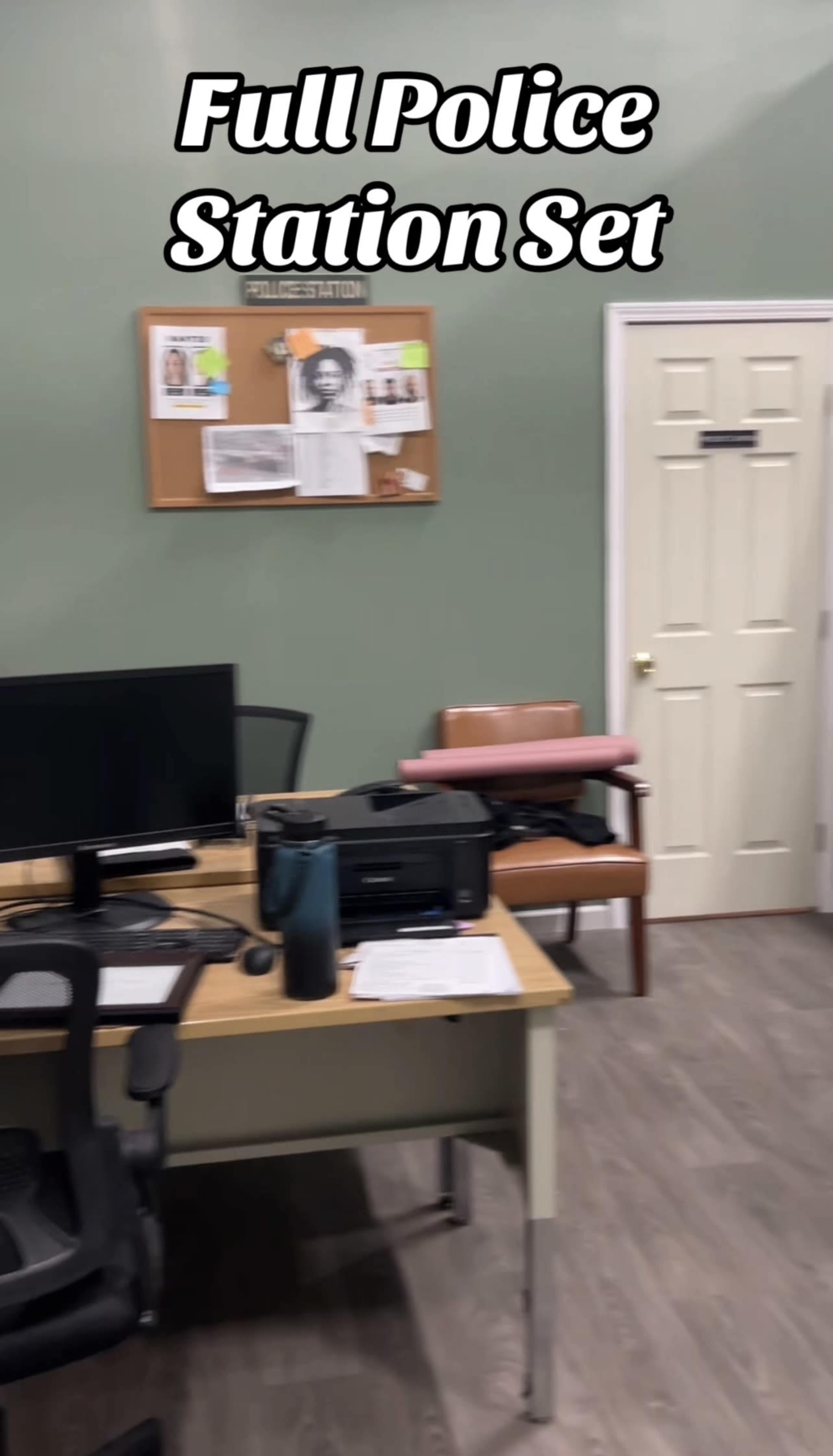 The image depicts an interior of a police station set, featuring a desk with a computer, a bulletin board on the wall, and a door leading to another room.