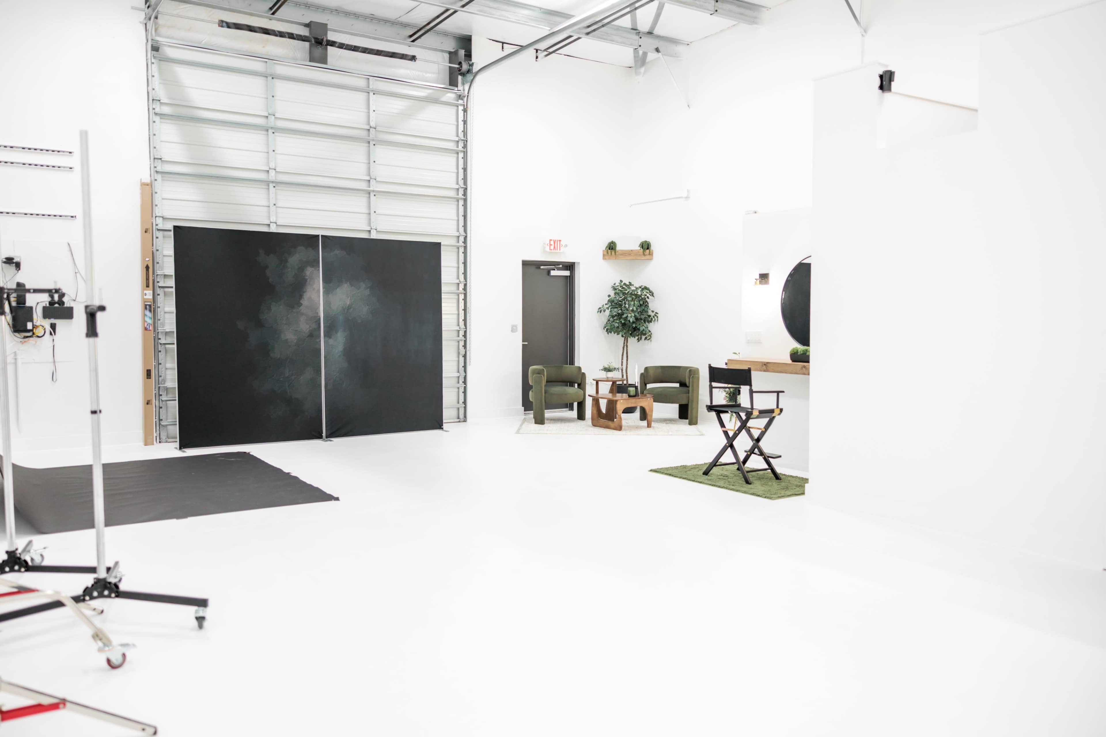 The image shows a bright, spacious studio with a black backdrop, a small seating area with green chairs, and minimalist decor.