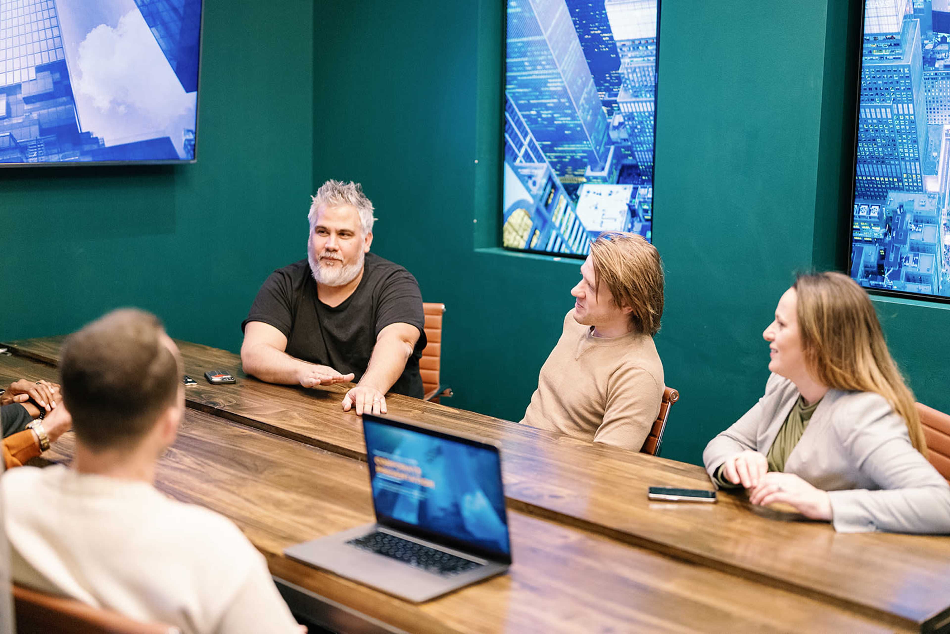 A group of four people is engaged in a discussion around a conference table in a room with large windows displaying an urban landscape.