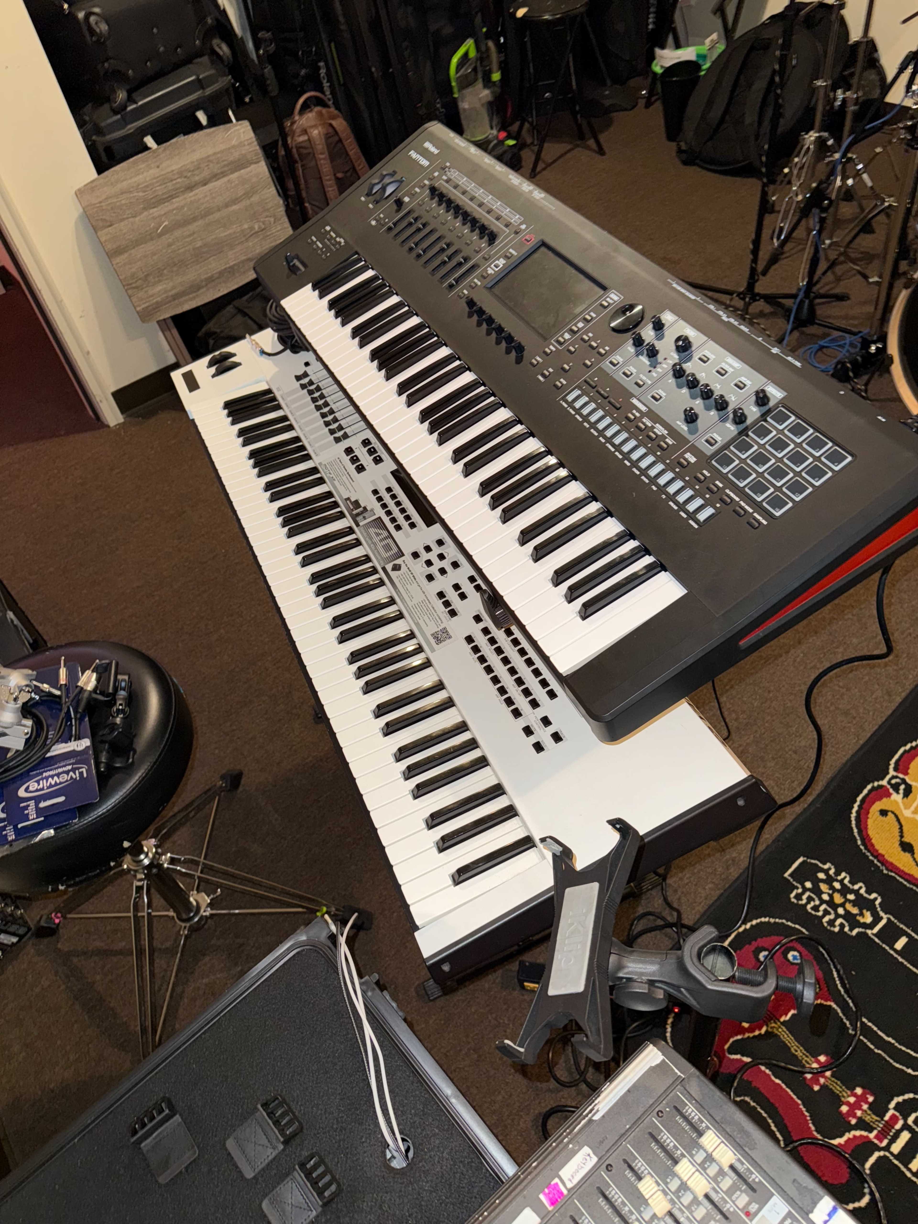 The image shows two electronic keyboards set up in a music rehearsal space, with drums and music equipment in the background.