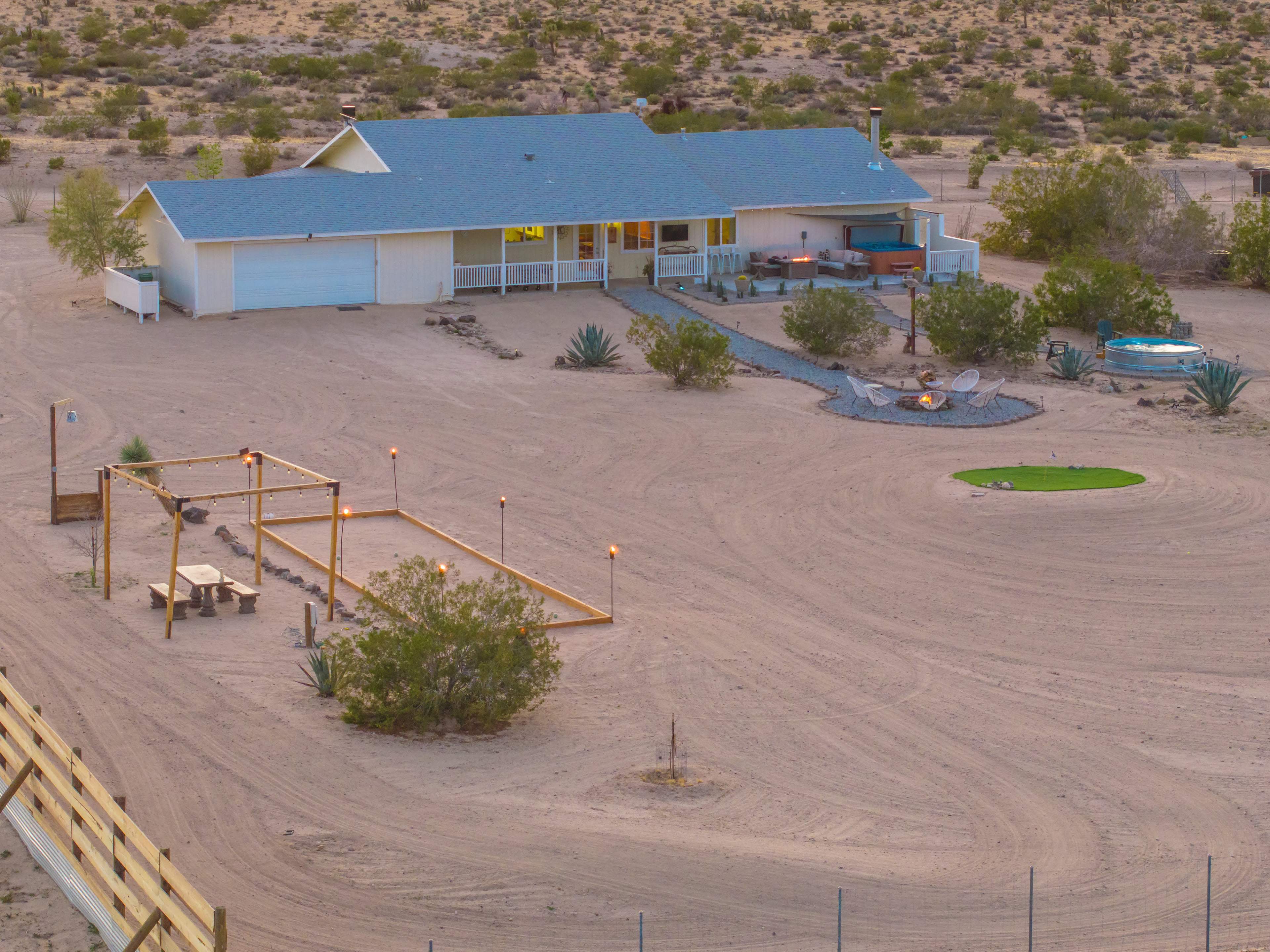 A single-story house with a blue roof is surrounded by sparse desert vegetation and features a patio area, a fire pit, and a small green lawn.
