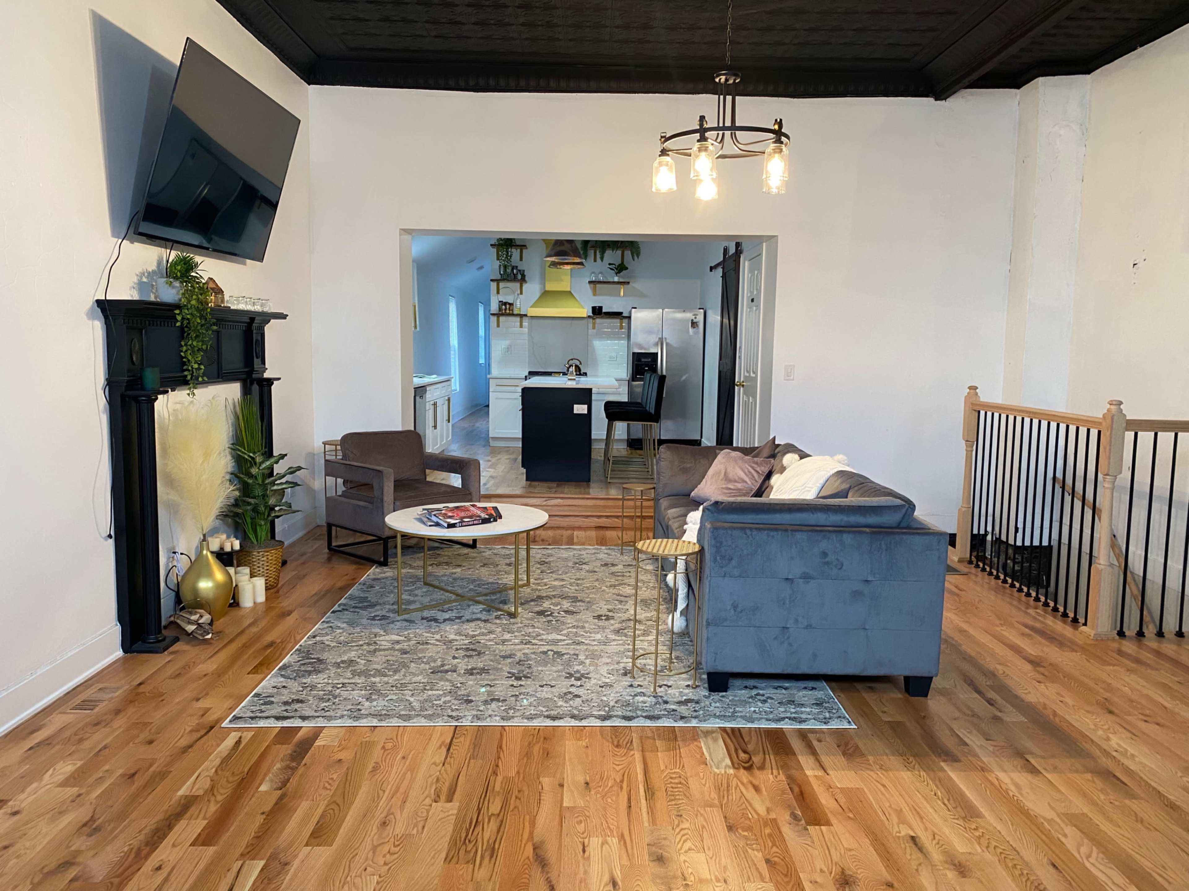 The living room features a gray sofa and chairs arranged around a coffee table, facing a wall-mounted television, with a view of the kitchen through an open space.