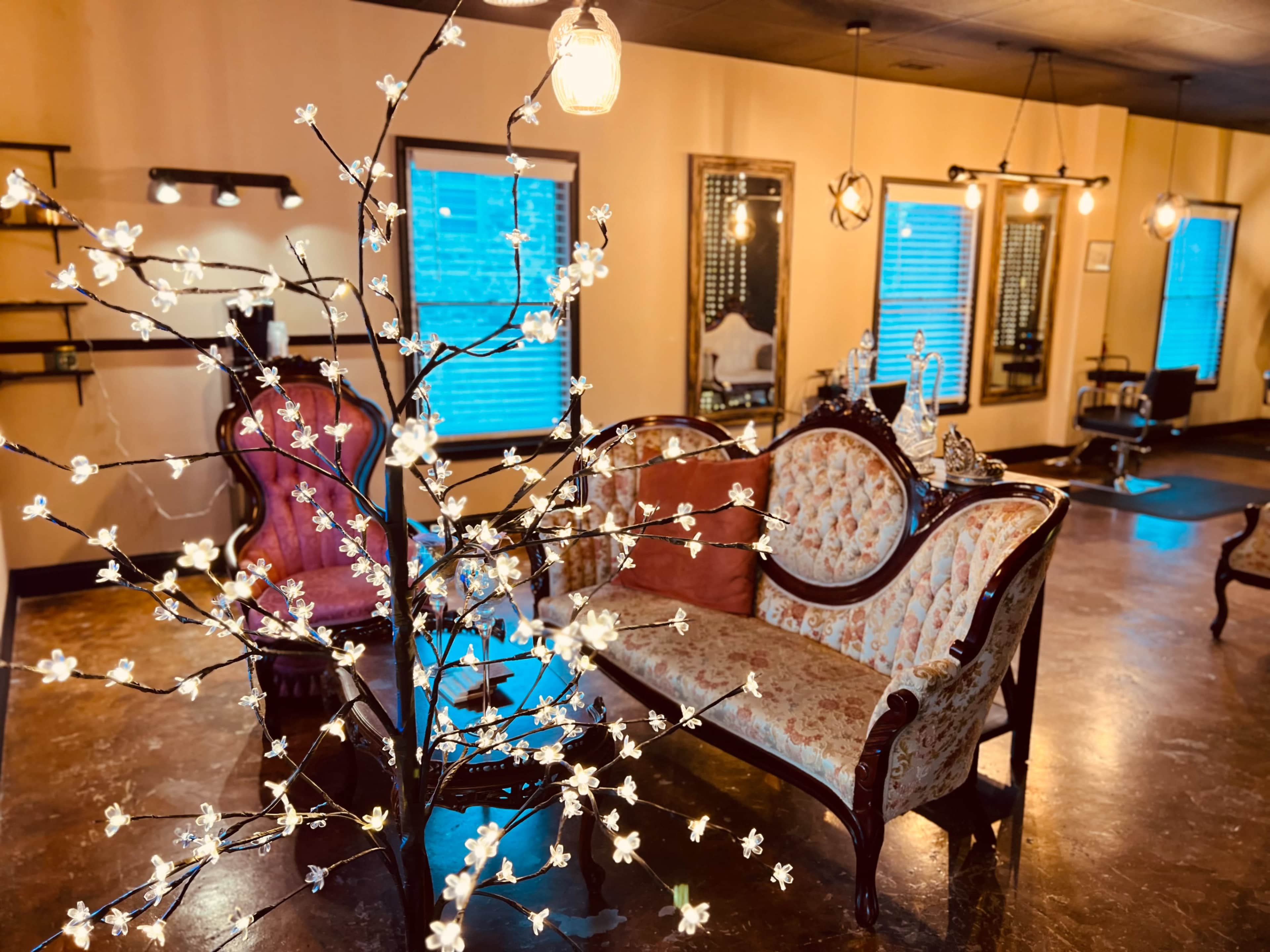 A vintage-style salon features a floral-decorated couch, a small table, and various decorative elements against a backdrop of softly lit walls and large windows.